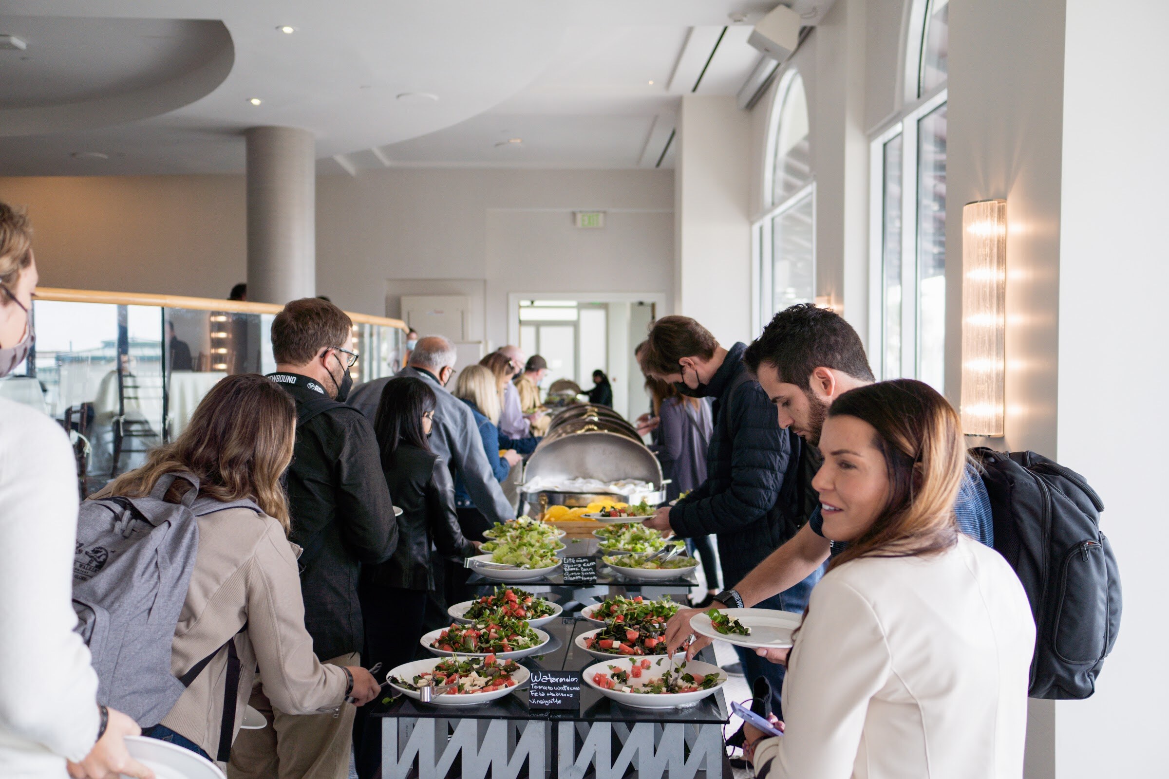 A line of attendees serving salads from a buffet table in a bright terrace room.