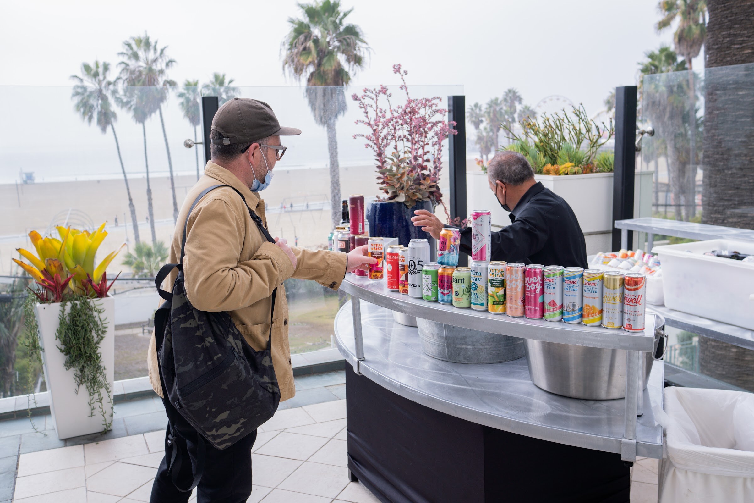 A man in a tan jacket and cap looks over a curved cart stocked with canned drinks on a terrace with palm trees and beach behind.