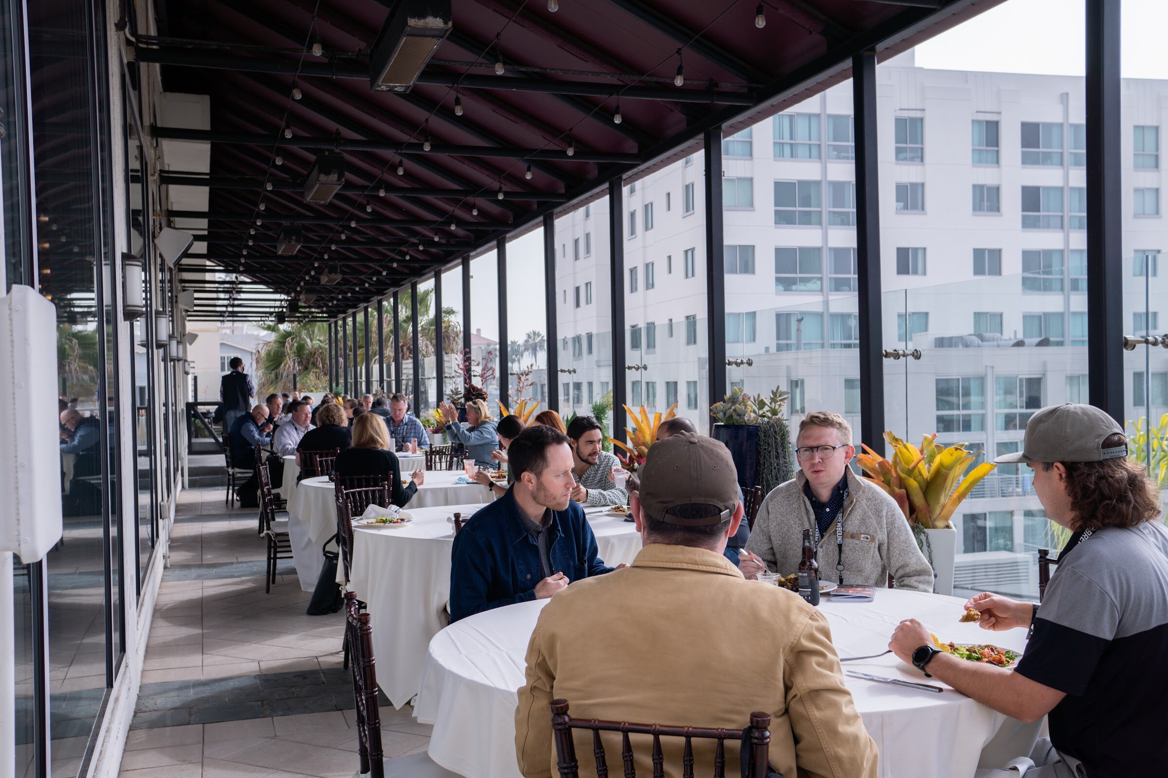 Attendees seated at round white tables along a long glass-walled terrace.