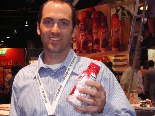 A smiling man in a striped shirt wearing a WhiteWave lanyard holds a red bottle. Behind him is a Silk Live display of bottles.