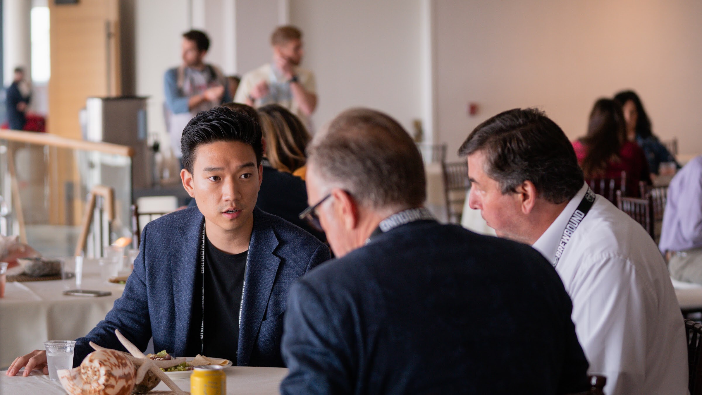 Three men in lanyards lean toward each other at a lunch table, the one at left looking serious.