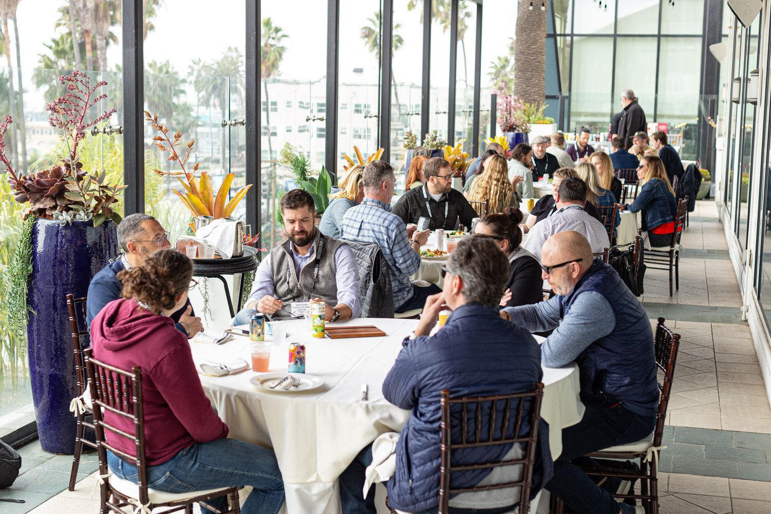 A long terrace lined with round white tables of attendees talking, with palm trees and tall windows behind.