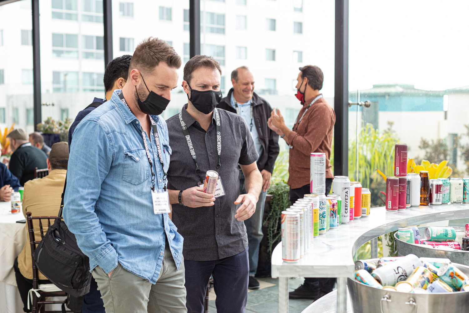 Two men in lanyards and masks stand at a curved metal cart lined with cans and ice buckets on a terrace.