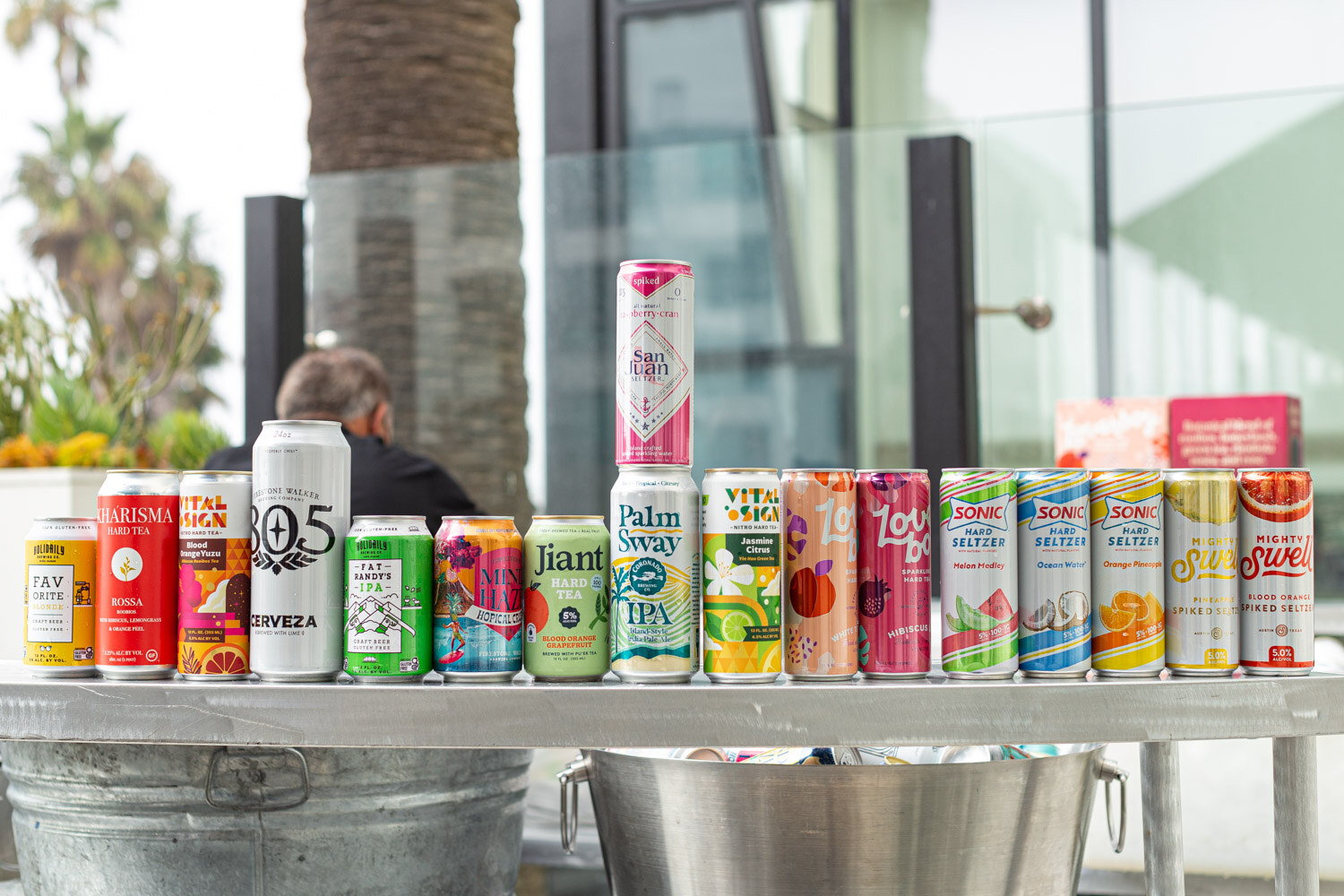 A row of colorful beverage cans, including Kharisma, 805 Cerveza, Palm Sway and Sonic, lined up on a metal cart with a bucket below.