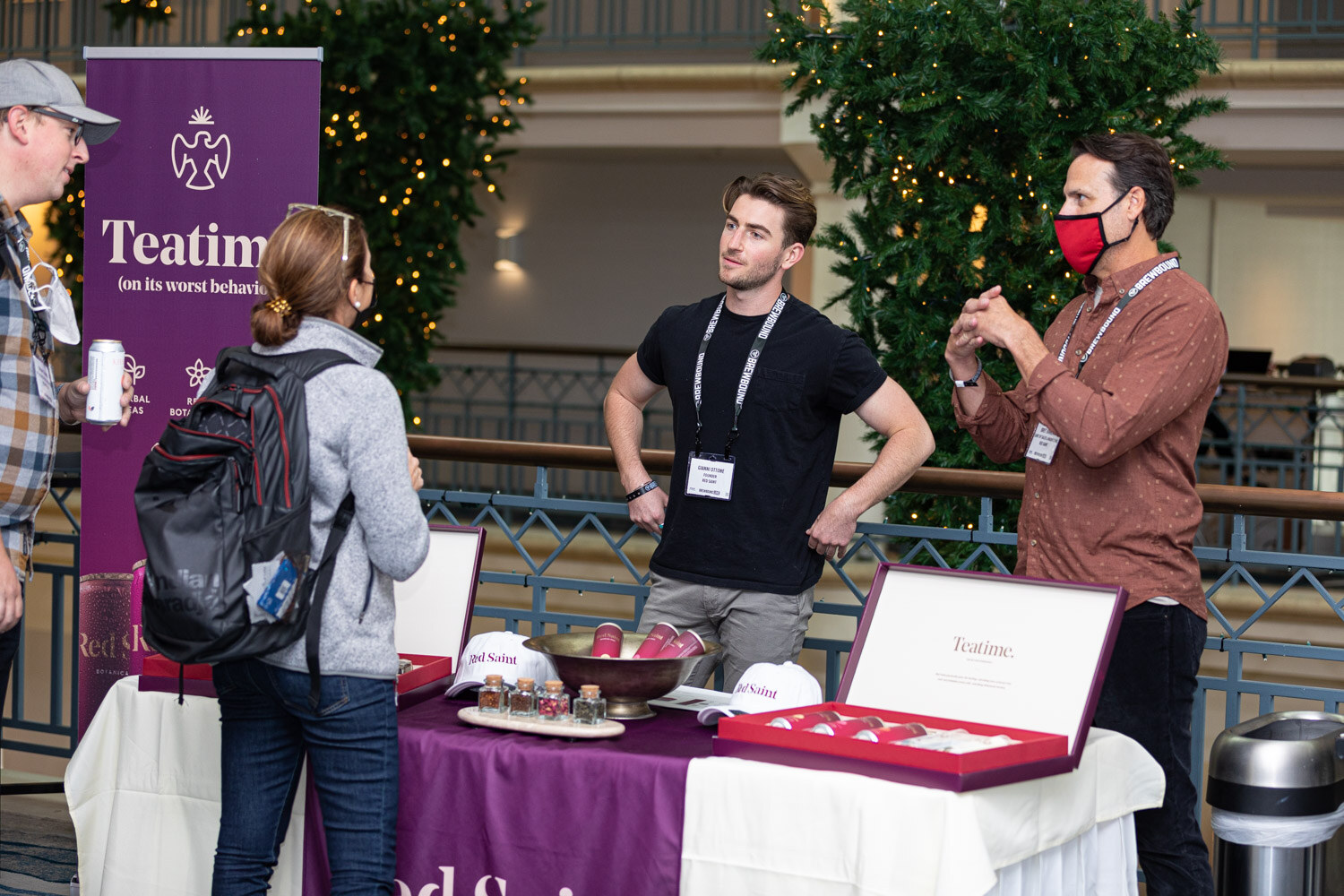 A table with a purple Teatime banner and boxed tea samples, with two men and a woman in a backpack standing in a hotel atrium.