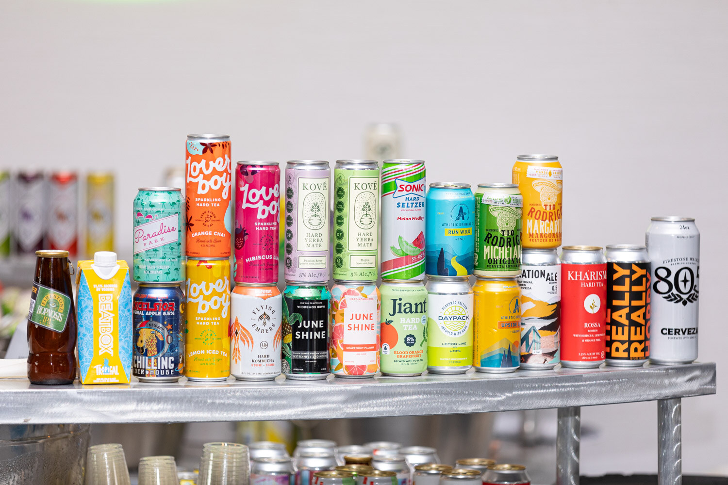 Two shelves of colorful cans and a few bottles, including Love Boy, Kove, JuneShine, Jiant and Kharisma, on a metal cart.