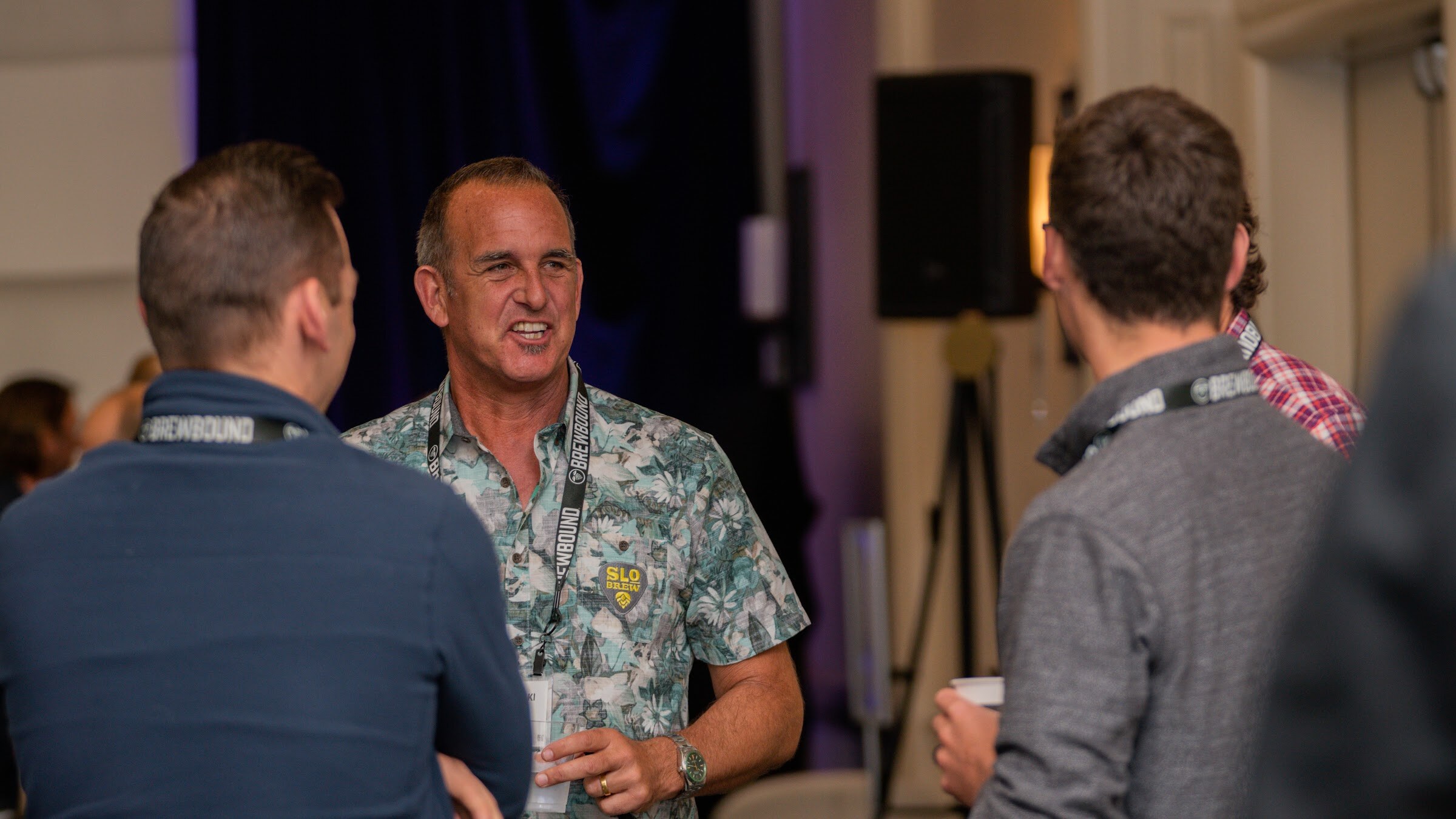 A man in a floral shirt and lanyard smiles while talking with two men facing away from the camera.