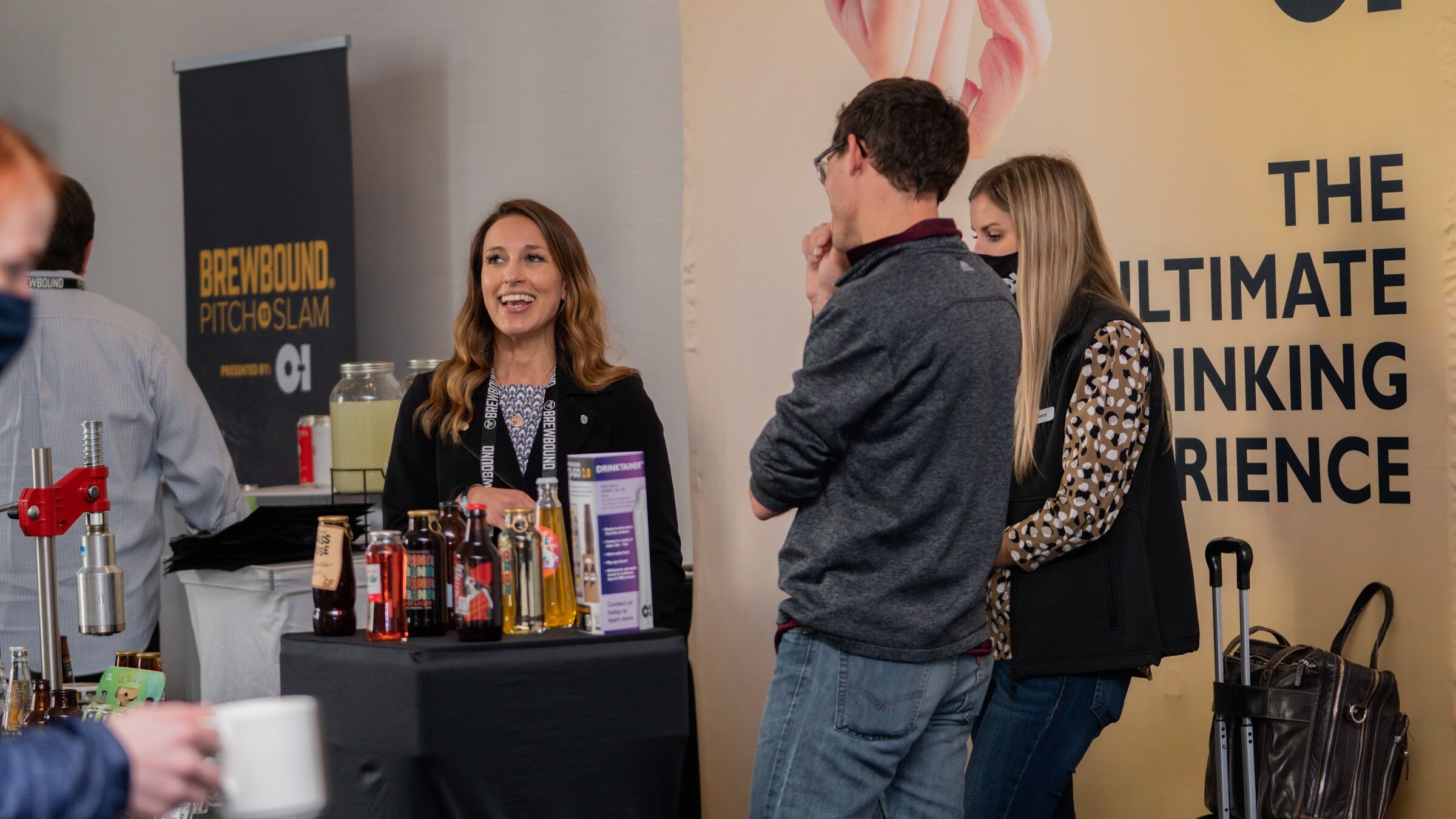 A black table with bottles and a small bar tool beside a Brewbound Pitch Slam banner and an O-I banner, with a smiling woman and two other people.
