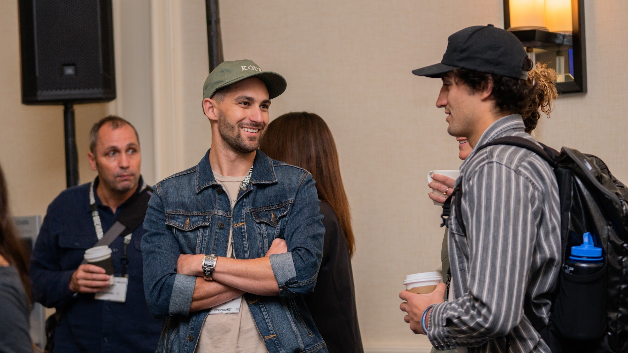 A smiling man in a Kove cap and denim jacket with crossed arms stands next to a man in a striped shirt with a backpack.