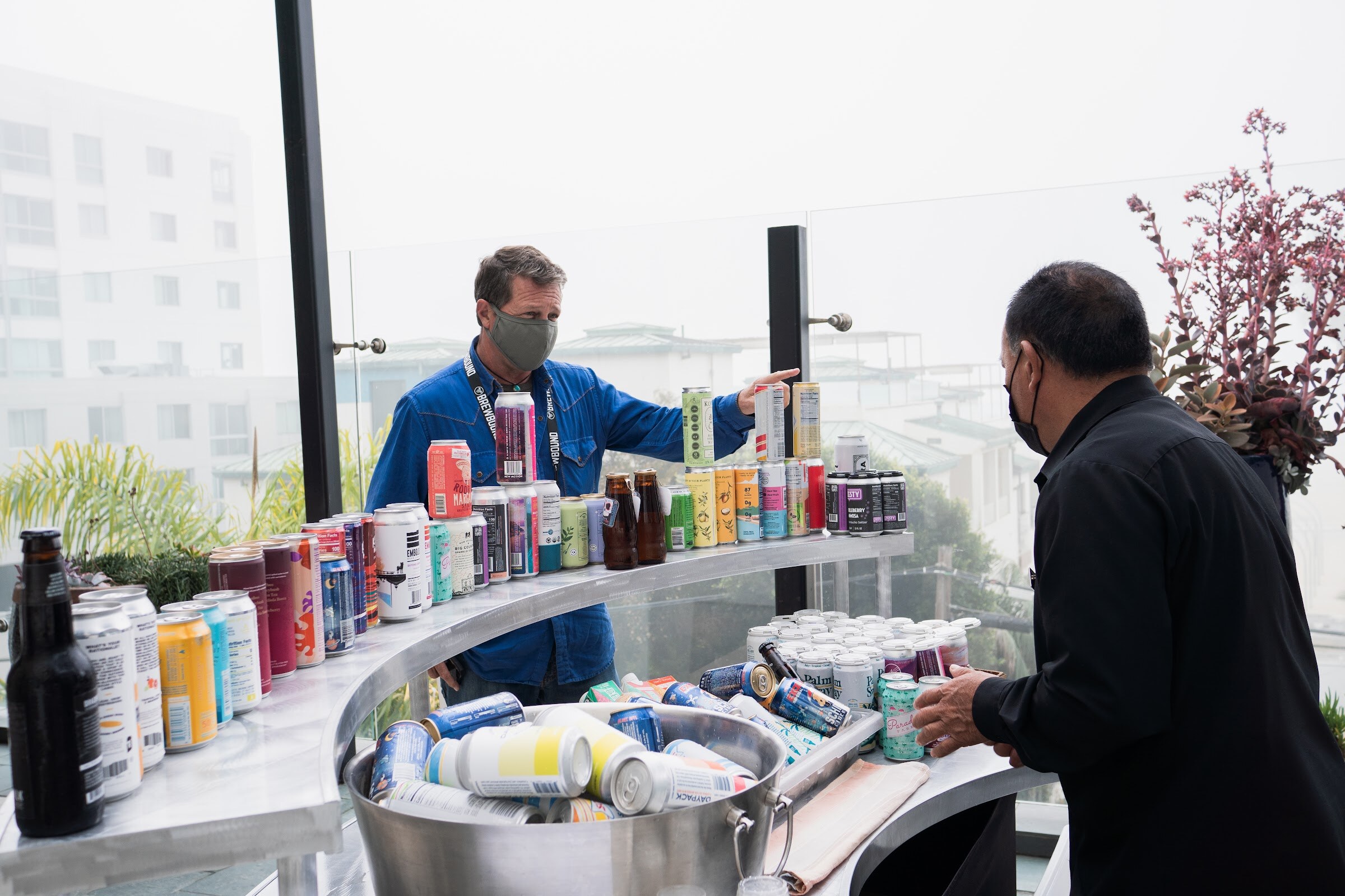 A man in a blue shirt and mask arranges cans on a curved cart while another man in black reaches in, with a coastal view behind.