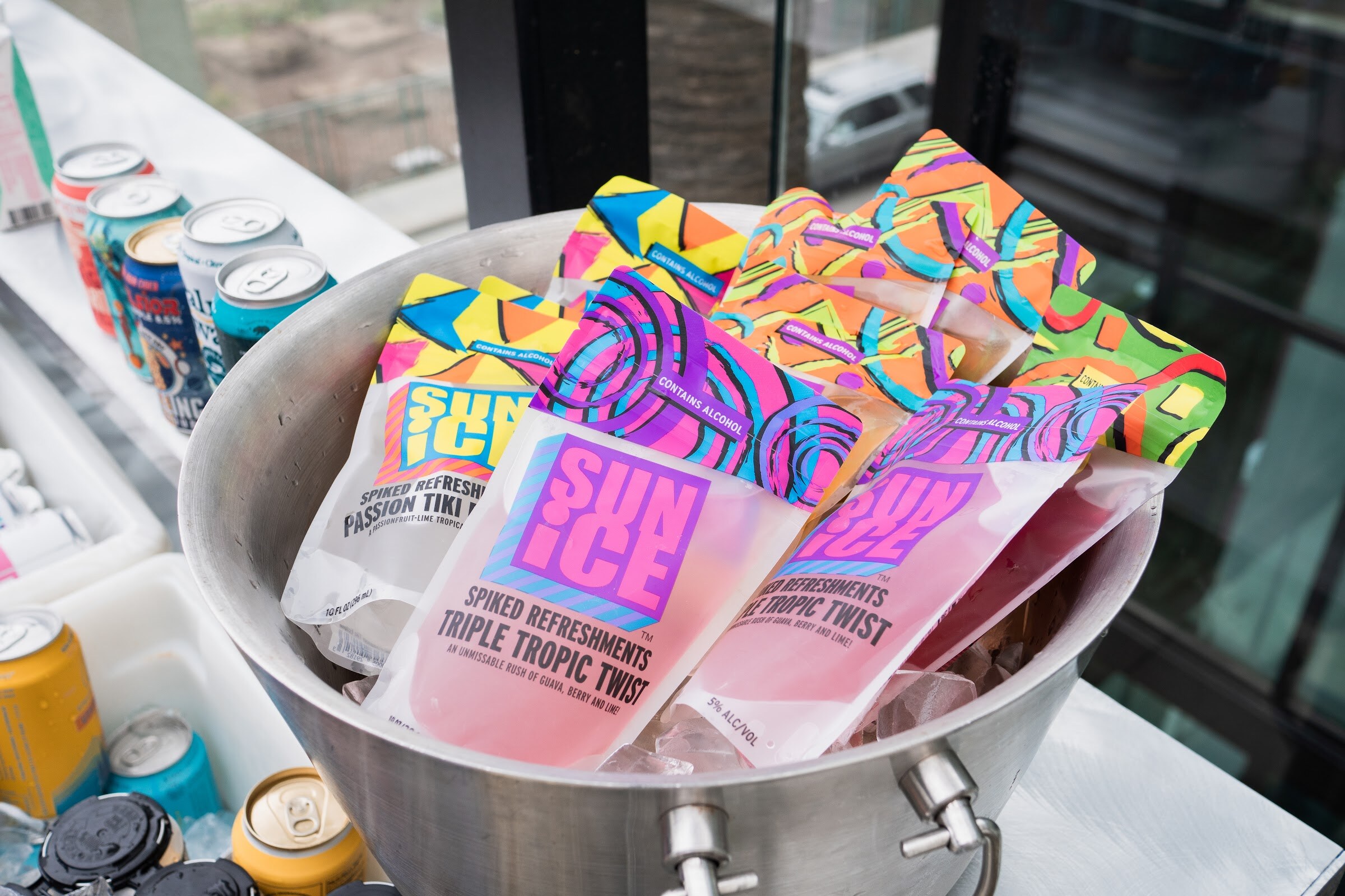 A metal bucket of colorful Sun Ice Spiked Refreshments pouches, Passion Tiki and Triple Tropic Twist, with cans beside it.