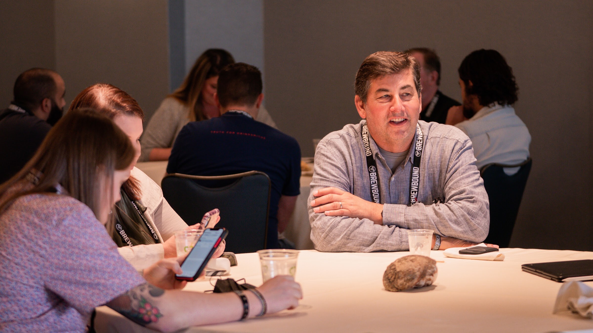 A smiling man in a gray shirt and lanyard sits at a white-clothed round table with other attendees, one looking at a phone.