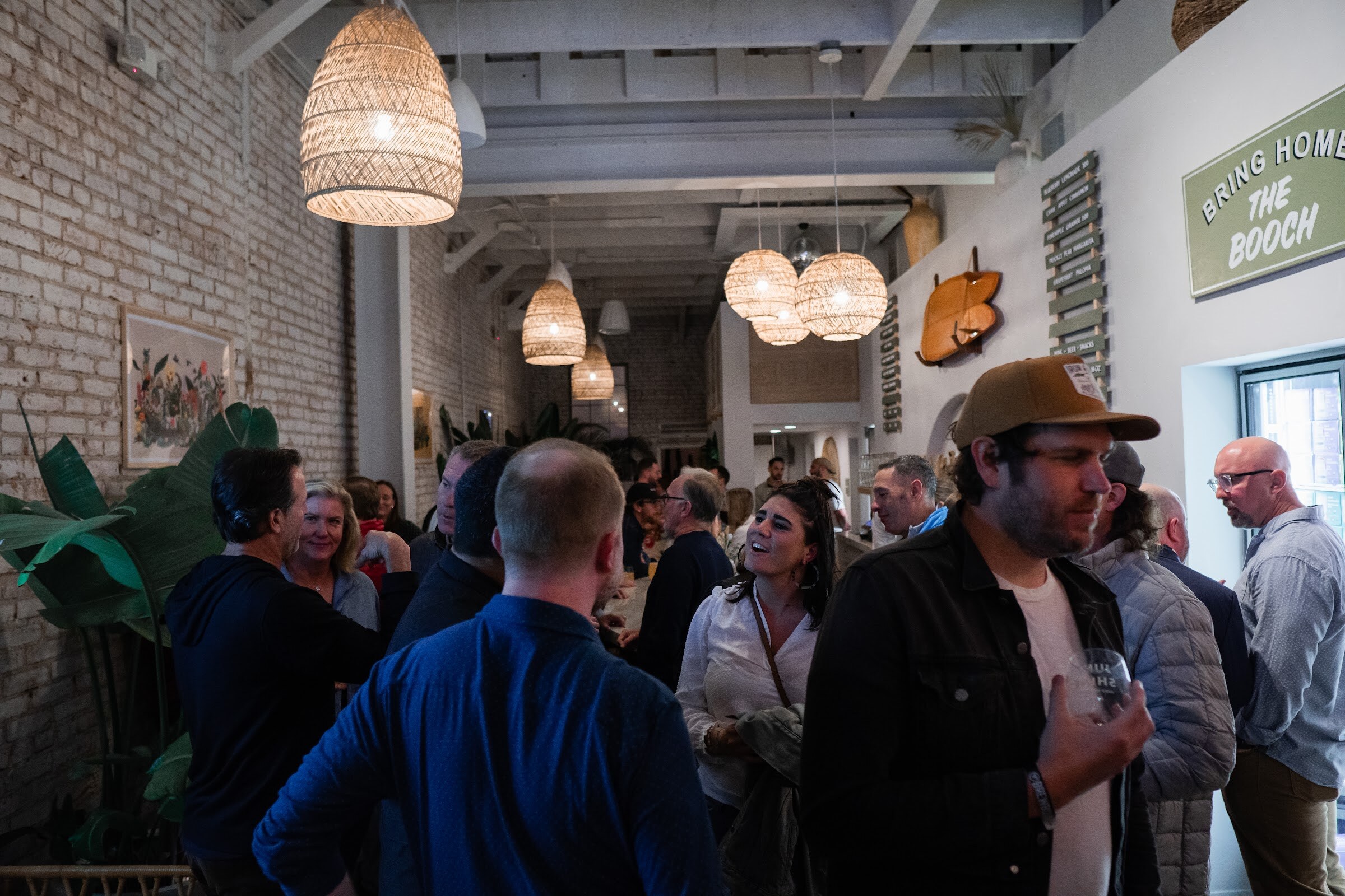 A crowd of people talks inside a brick-walled taproom with woven pendant lamps and a Bring Home the Booch sign.