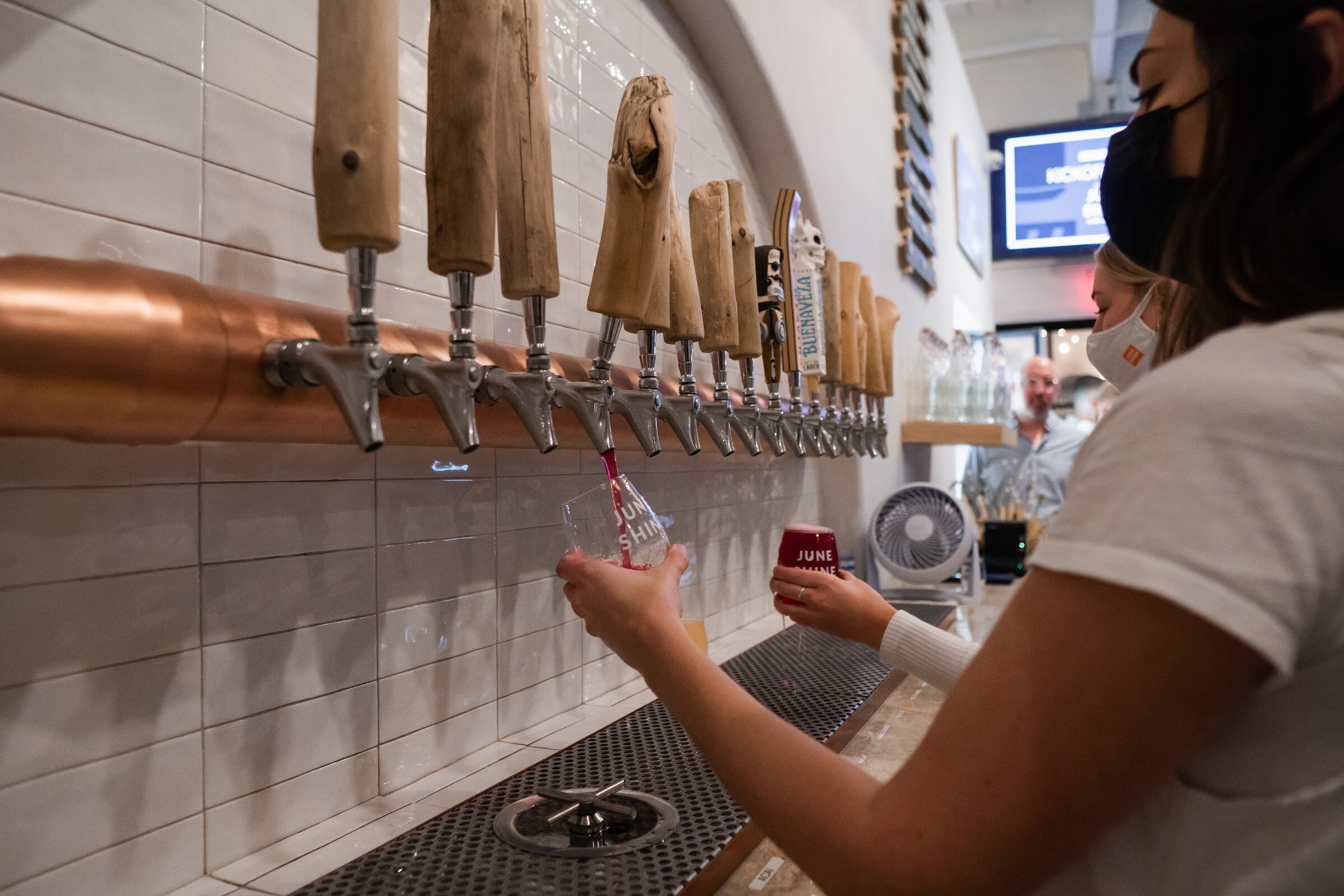 A woman in a mask pours drinks beneath a row of wooden tap handles over a copper rail.