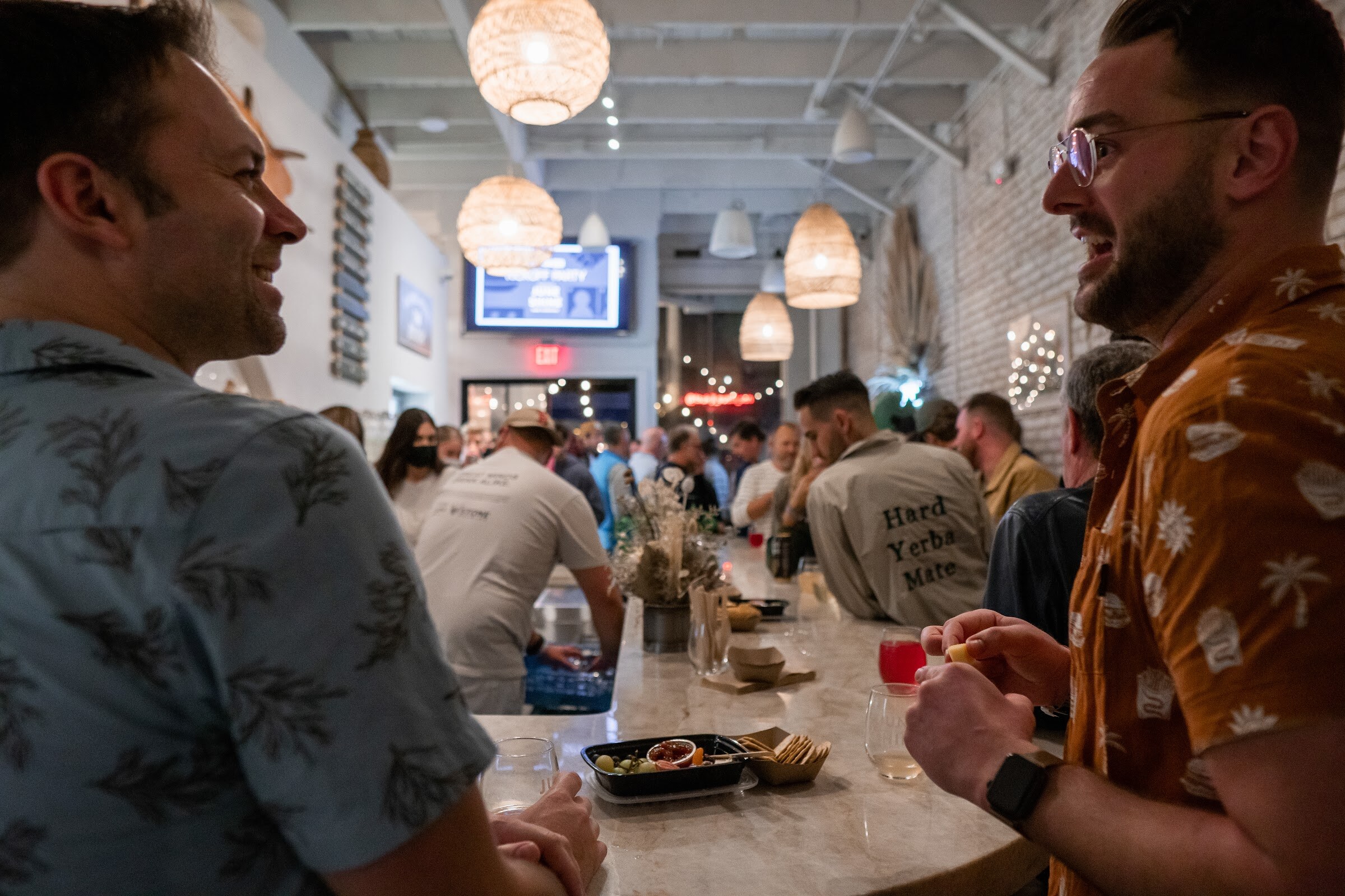 Two men in patterned shirts talk at a marble bar with snacks and drinks, with a crowd and TV screens behind them.