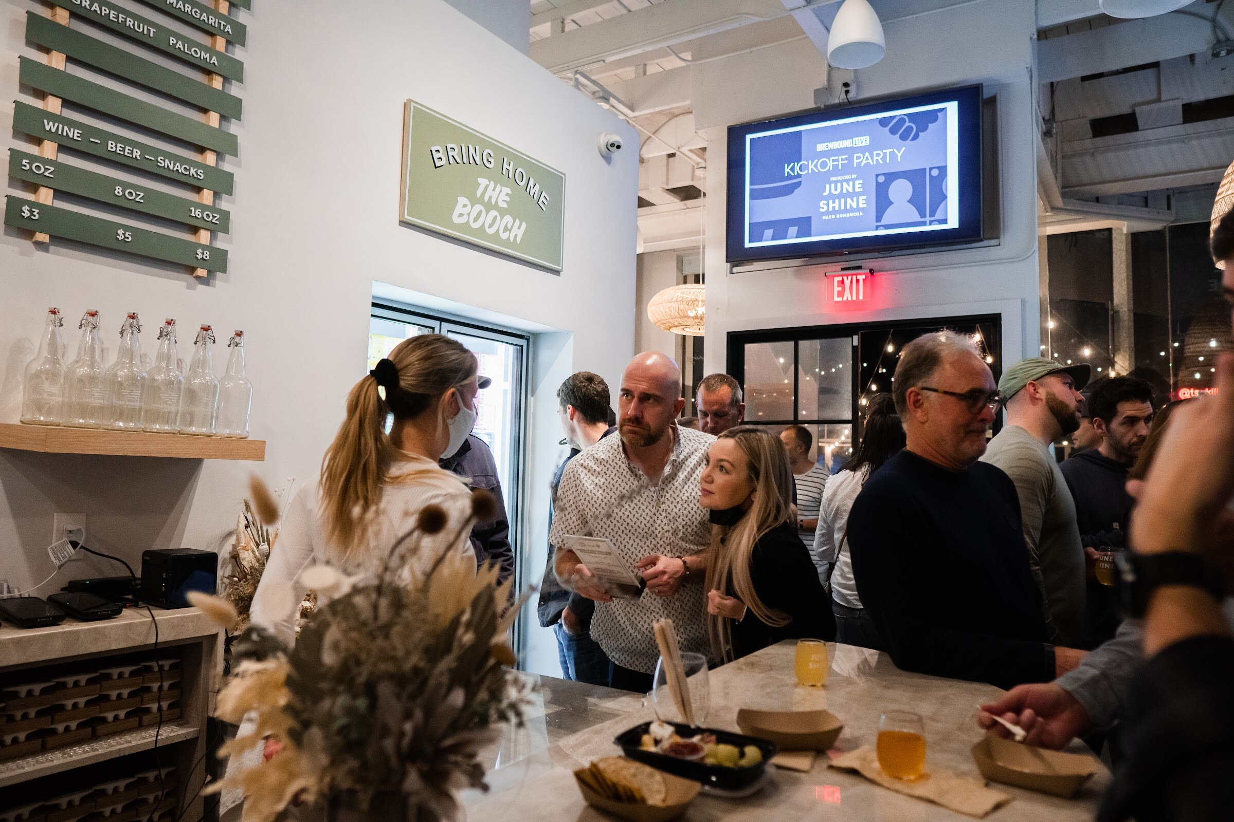 Guests stand around a marble bar with snack trays and drinks, a green sign reading Bring Home the Booch and a screen showing Kickoff Party behind them.