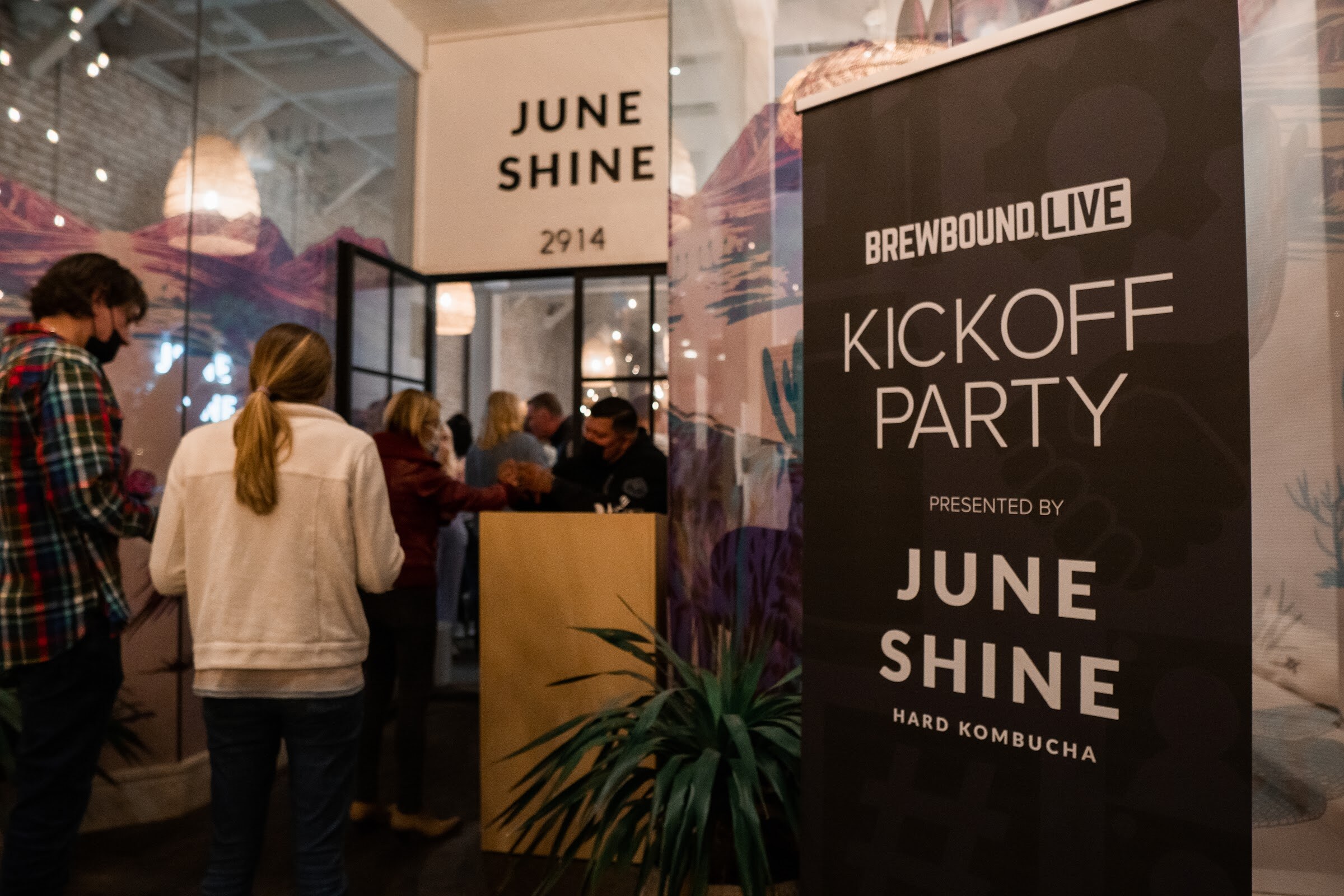 A tall black banner reading Brewbound Live Kickoff Party presented by JuneShine Hard Kombucha stands beside a JuneShine doorway with guests checking in.