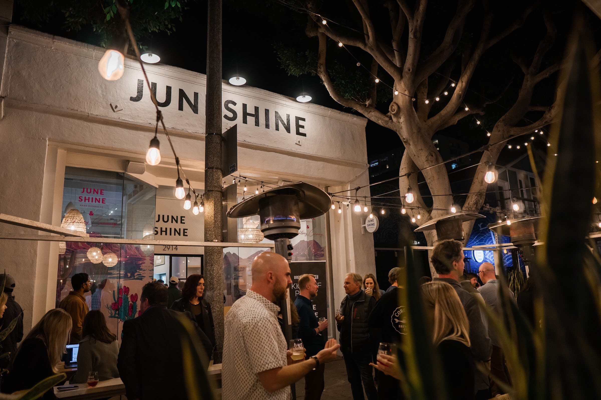 The JuneShine storefront at night with string lights across a tree-lined patio and guests talking with drinks.