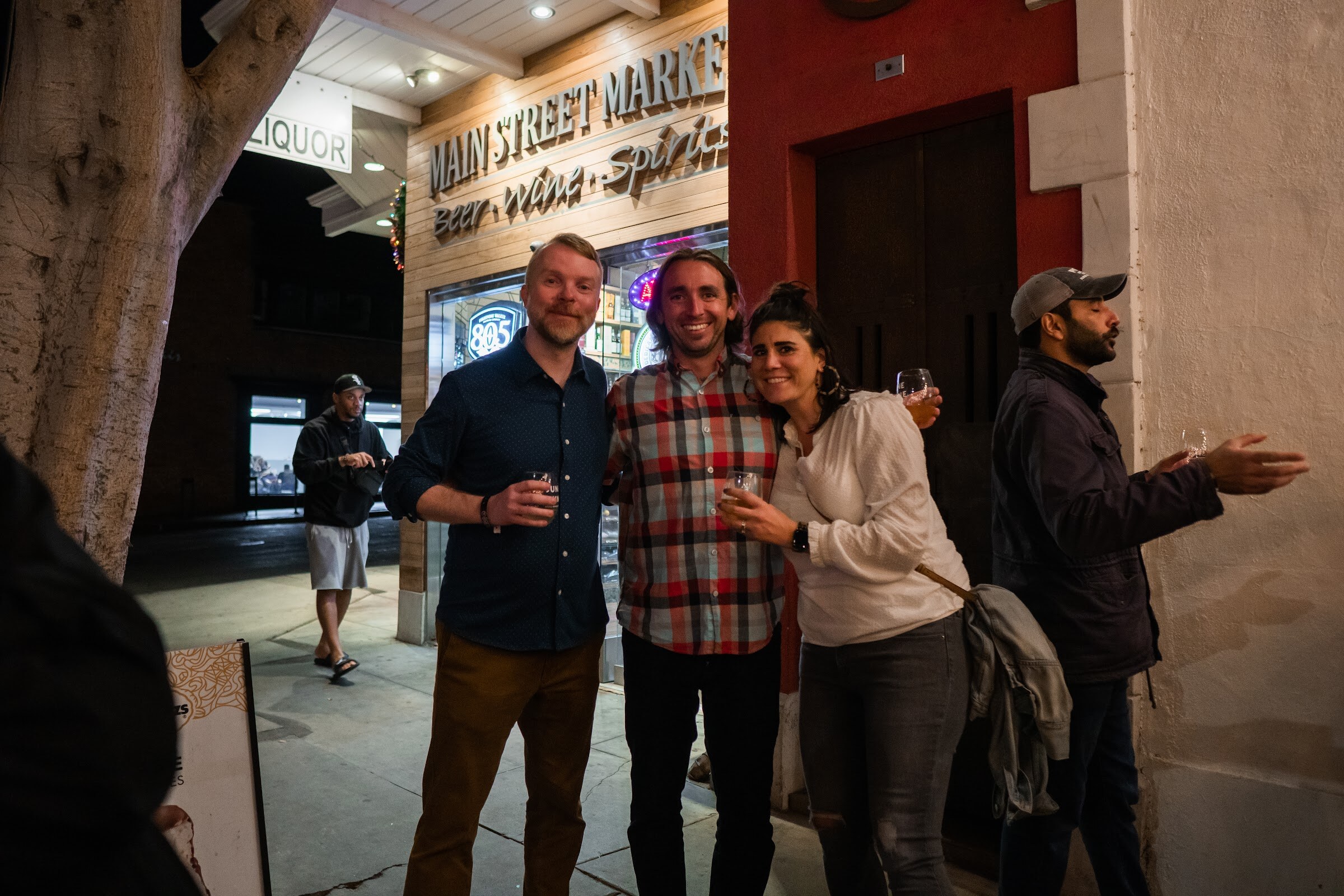 Two men and a woman hug and smile for the camera on a sidewalk in front of a Main Street Market sign at night.
