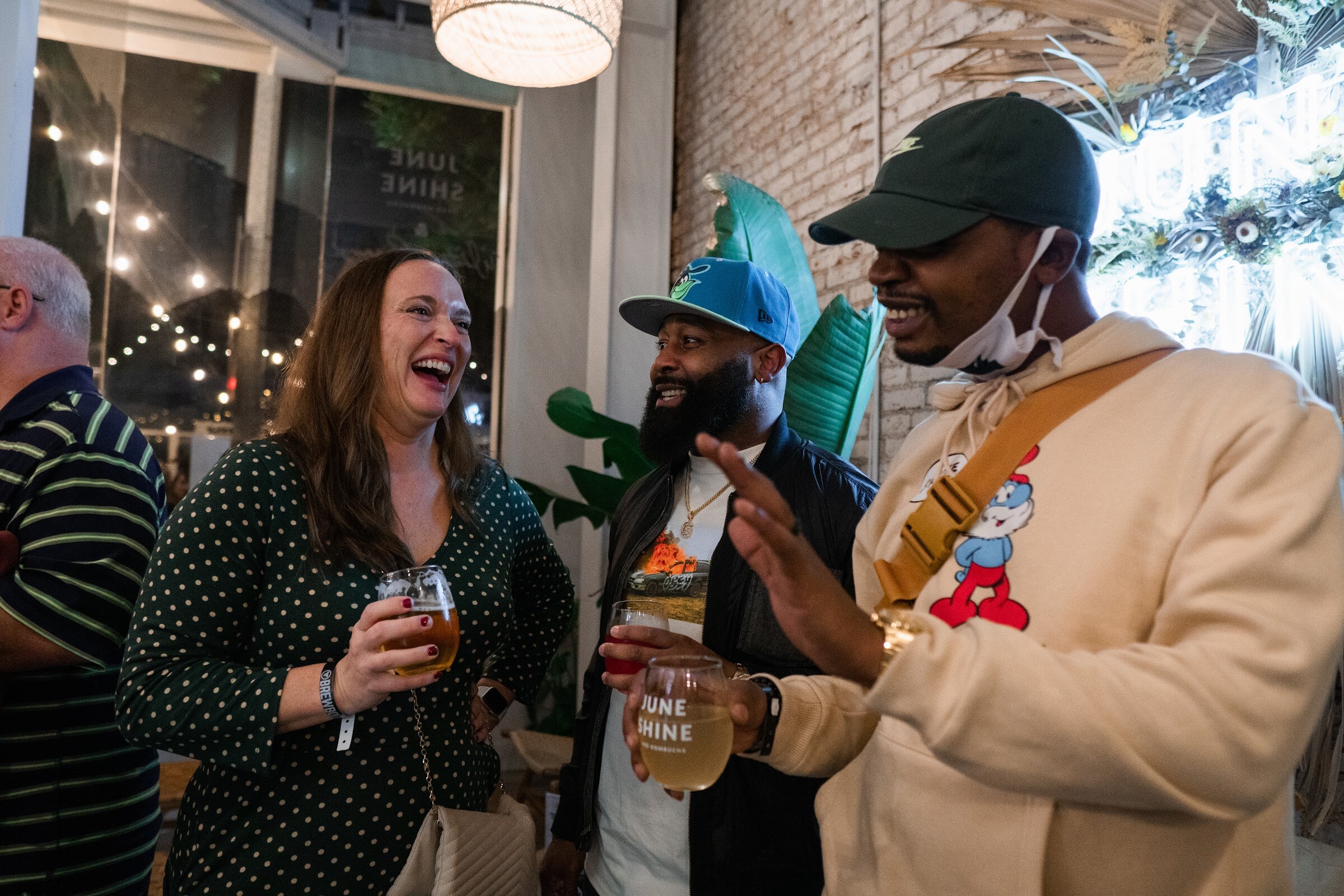 A woman in a polka-dot top laughs next to two men, one holding a JuneShine glass, inside the taproom.