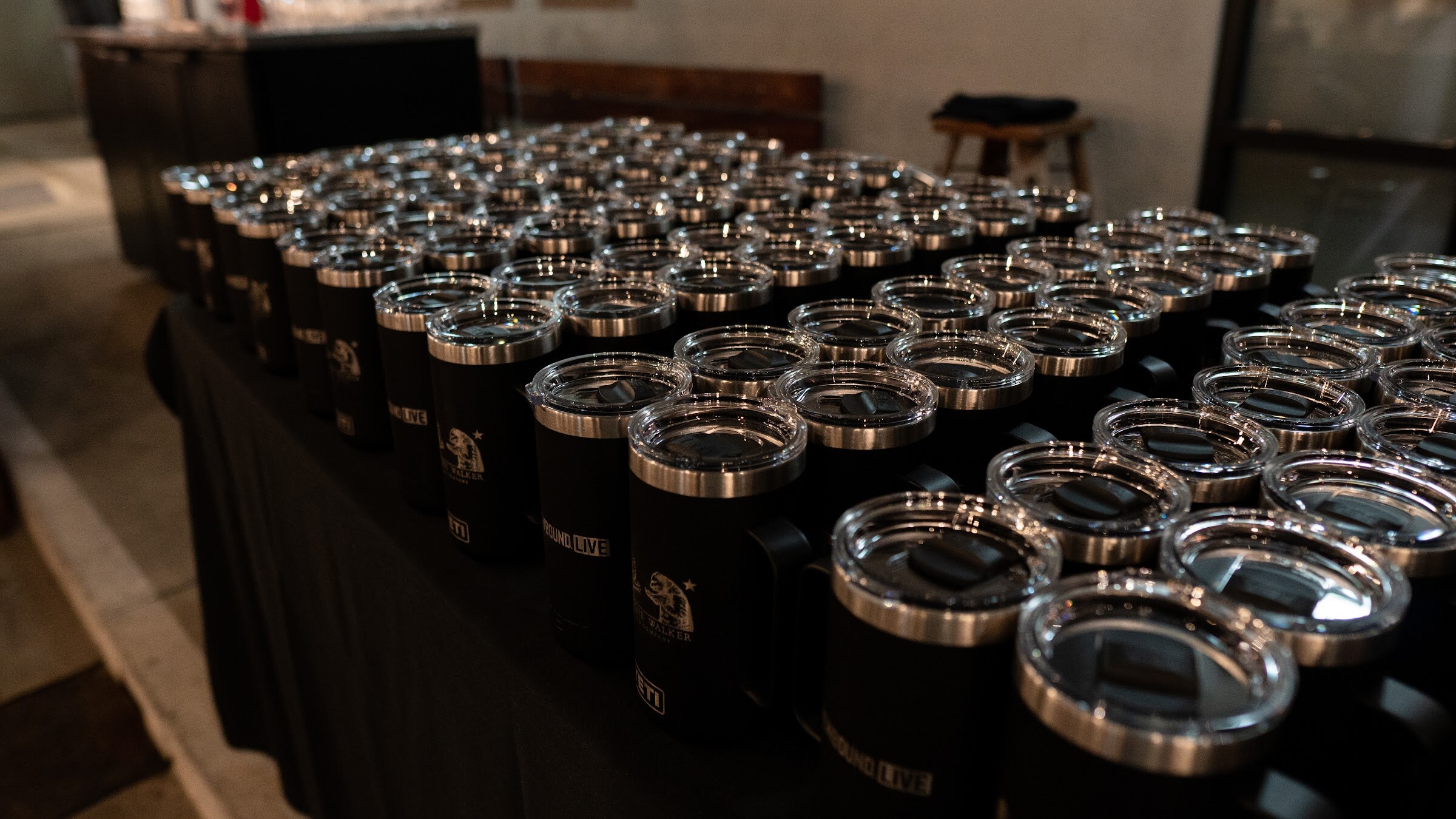Dozens of black insulated mugs with lids lined up on a table covered in black cloth.