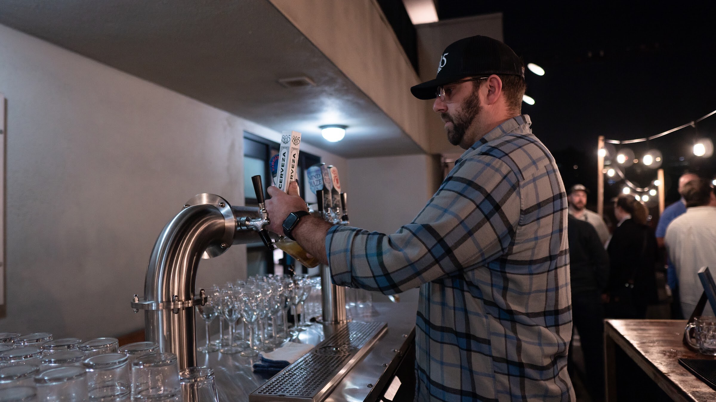 A bearded man in a plaid shirt and cap pulls a tap beside rows of glasses at an outdoor bar.