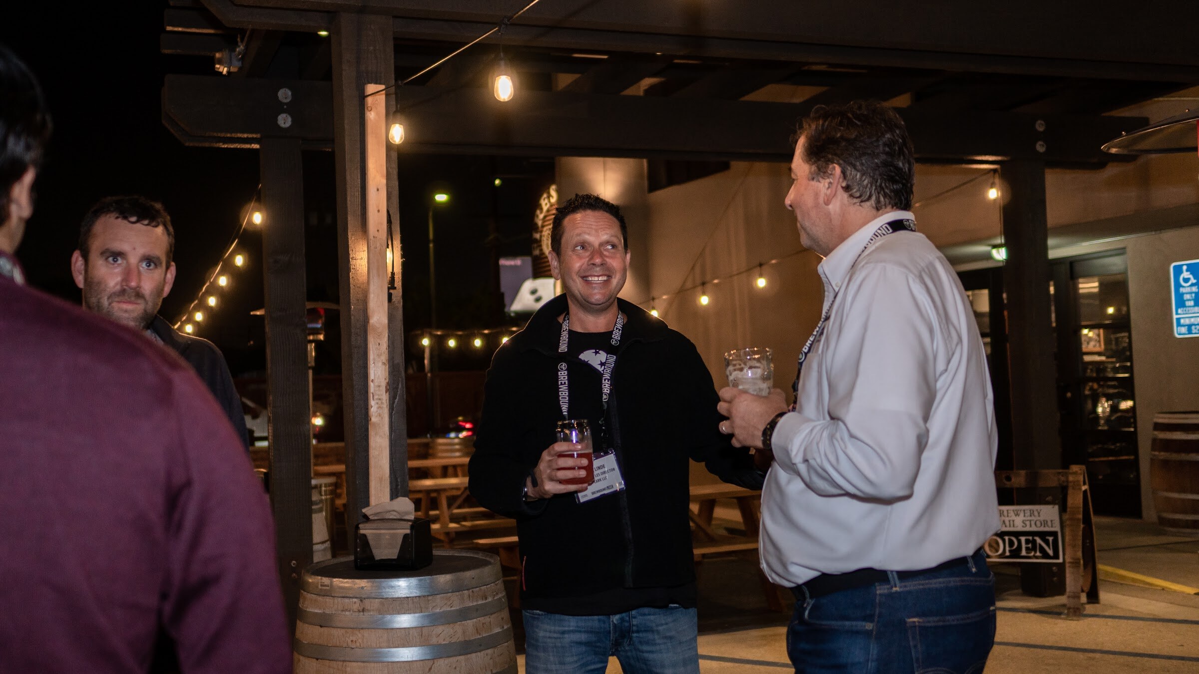 A smiling man in a dark jacket and lanyard holds a drink while talking with a man in a white shirt on a lit patio.