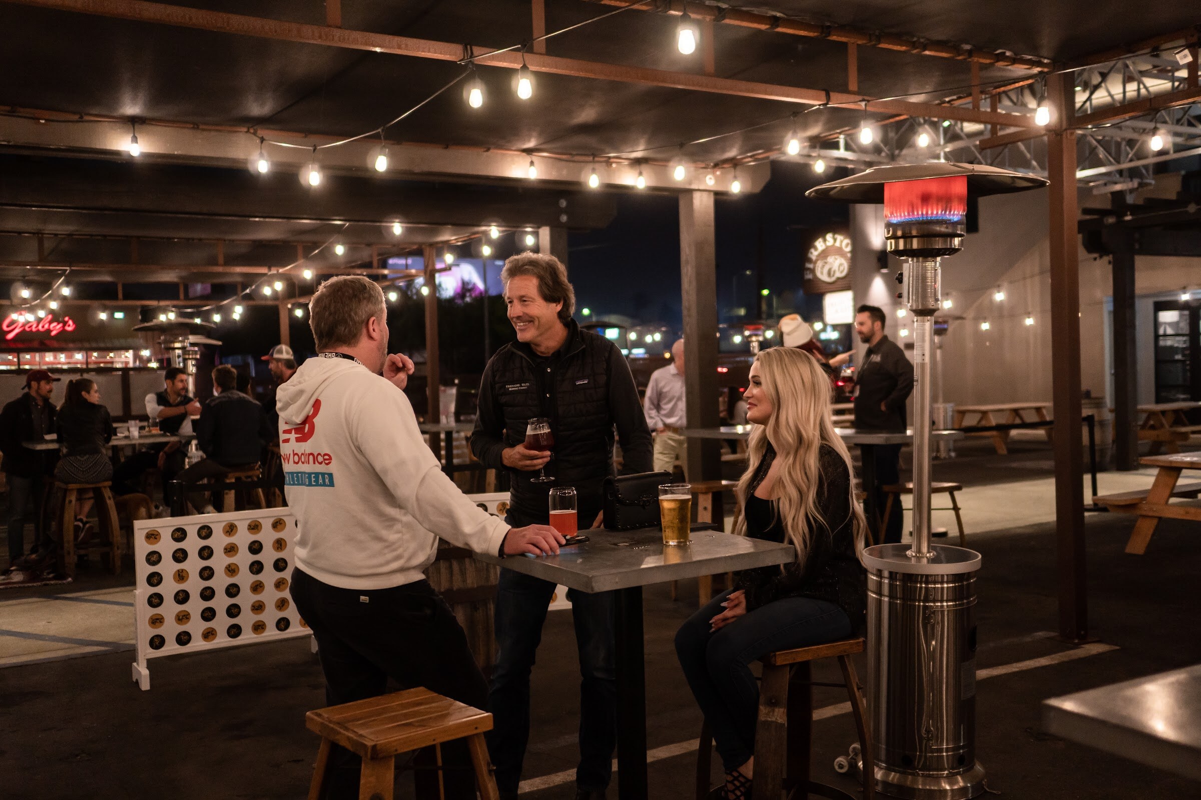Two men and a woman with beers stand and sit around a high table beside a giant Connect Four board under string lights.