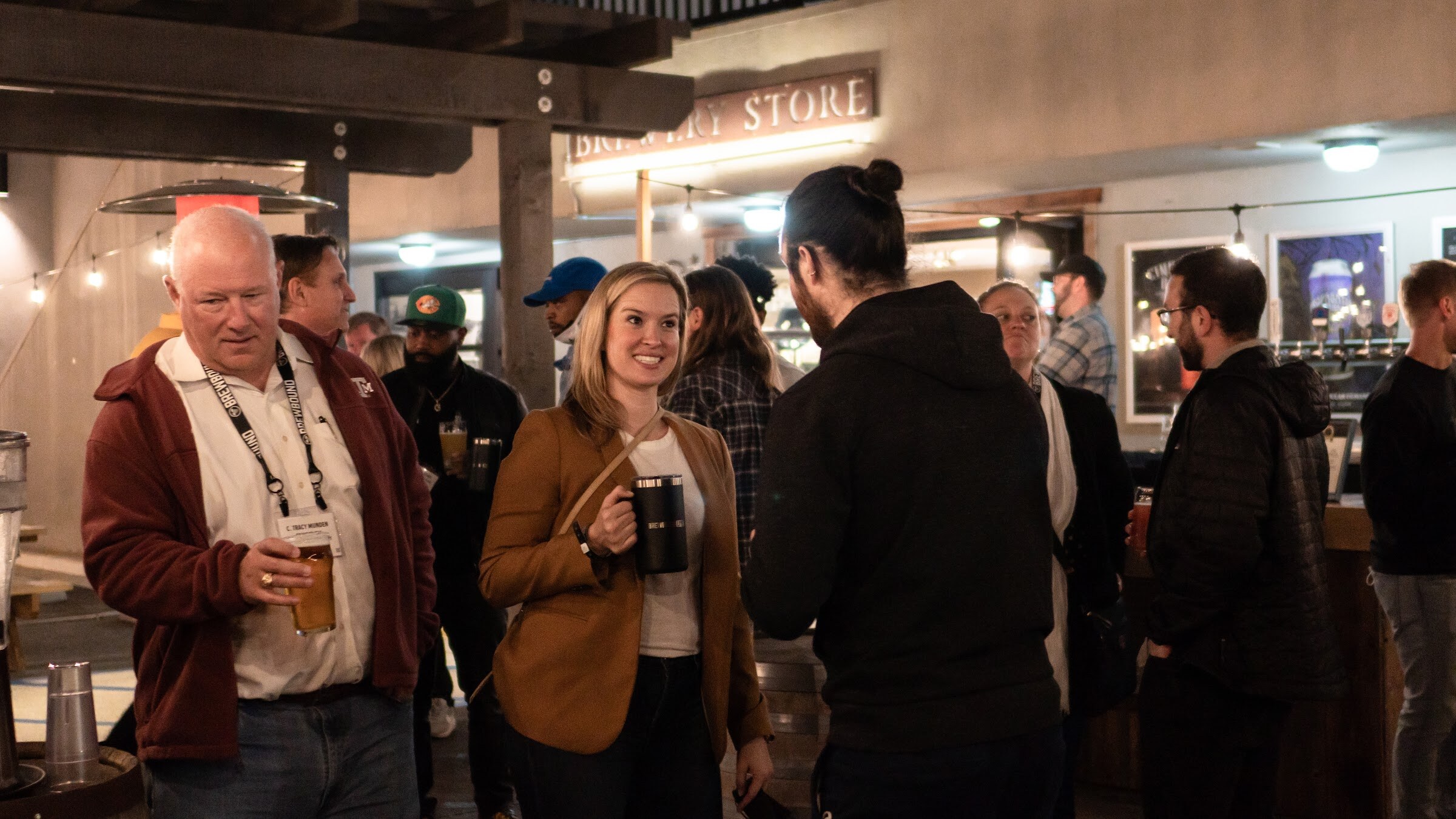 A man in a maroon jacket and a woman in a tan blazer smile and hold black mugs in a crowd outside the taproom.