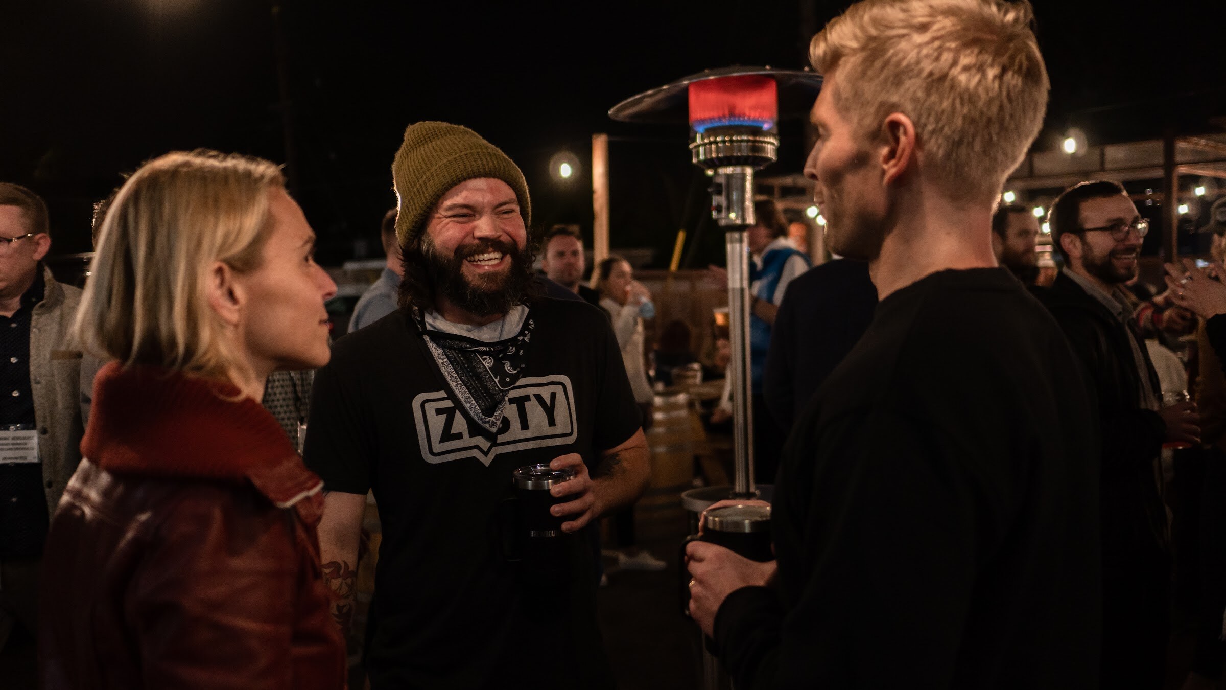 A man in a beanie and black Zesty t-shirt laughs while talking with a woman in a red jacket and a blond man.