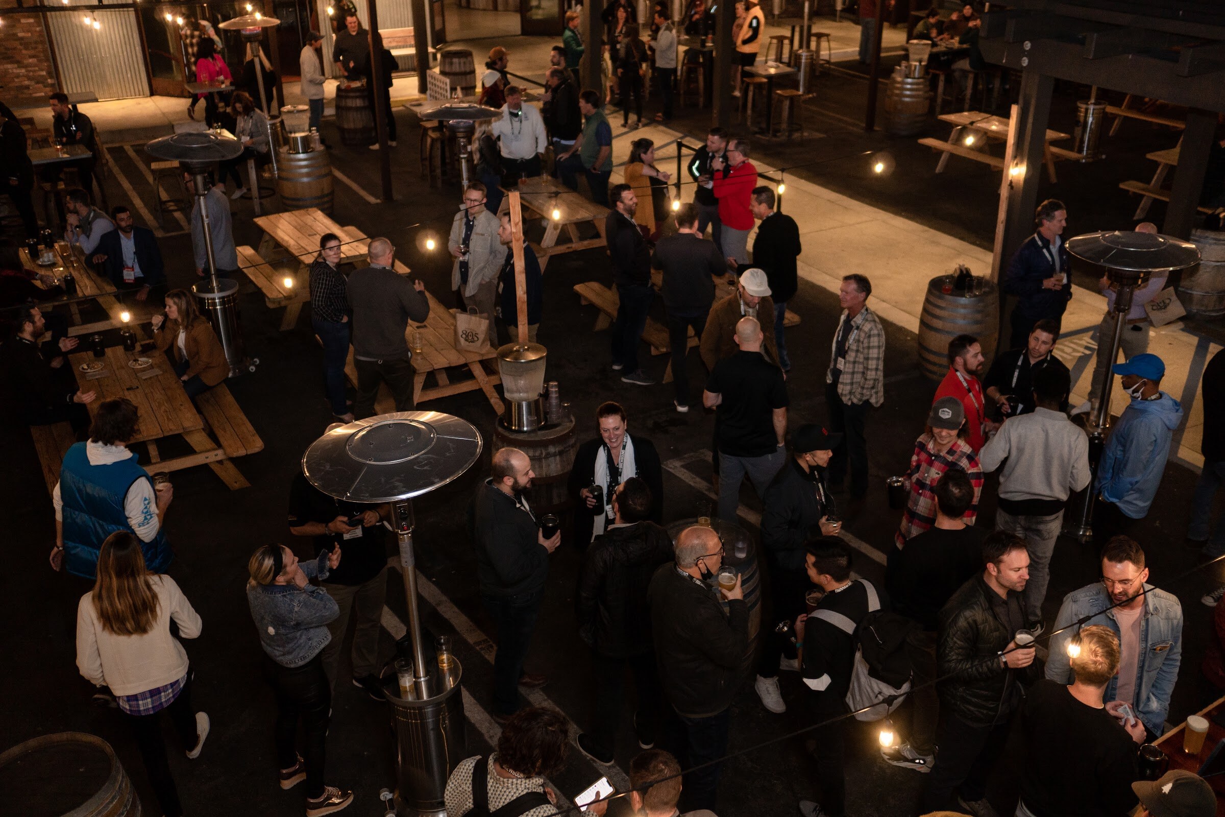 An elevated view of a crowded outdoor patio at night with picnic tables, patio heaters and string lights.