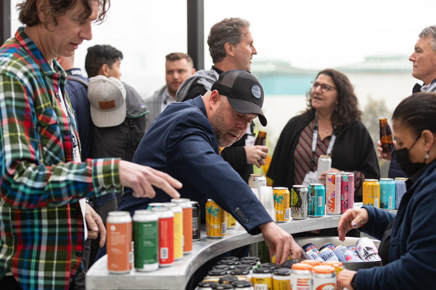 A group of attendees crowd around a curved metal cart full of colorful cans, one man in a plaid shirt reaching in.