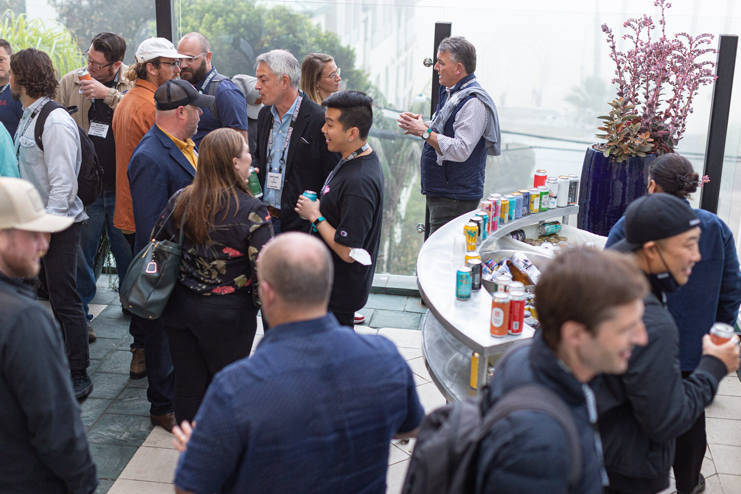 A crowd stands around a curved metal cart of cans on a terrace with a flowering plant in a blue pot.