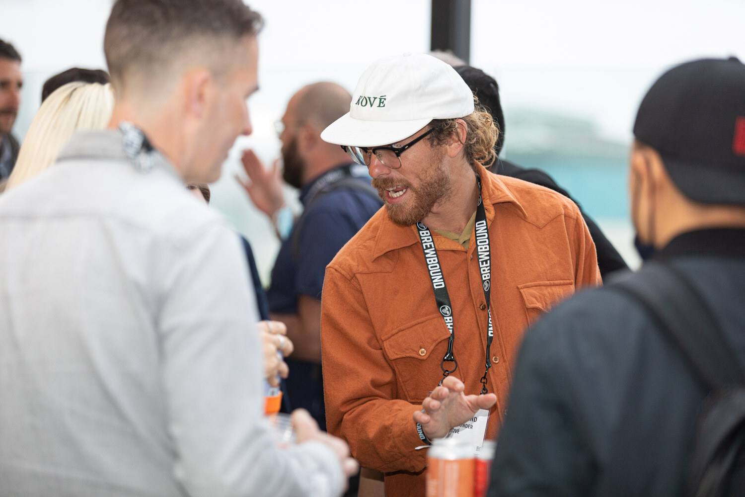 A bearded man in a white Kove cap, glasses and an orange shirt with a lanyard talks with people around him.