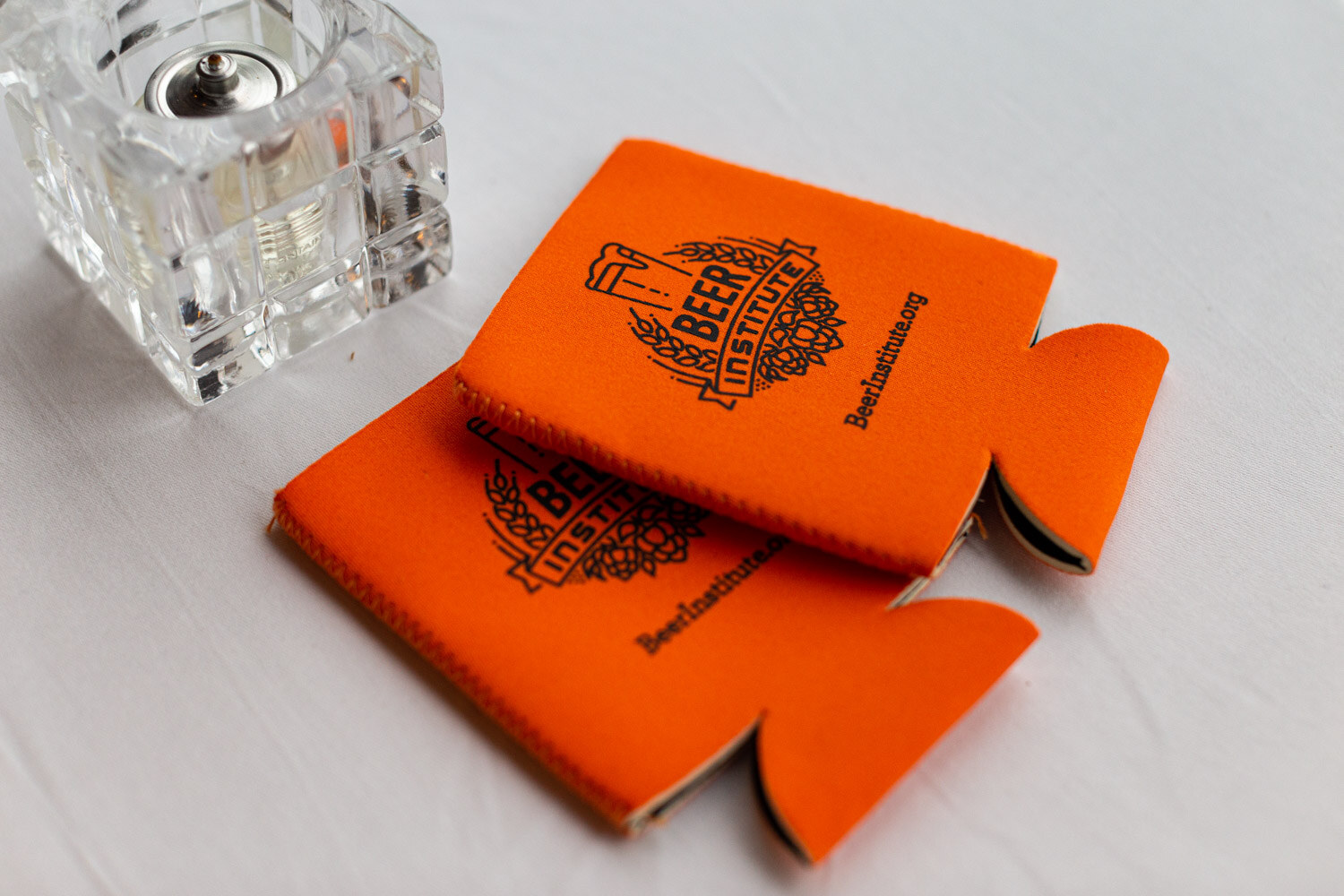 Two orange can coolers printed with the Beer Institute logo and beerinstitute.org on a white tablecloth beside a glass candle holder.