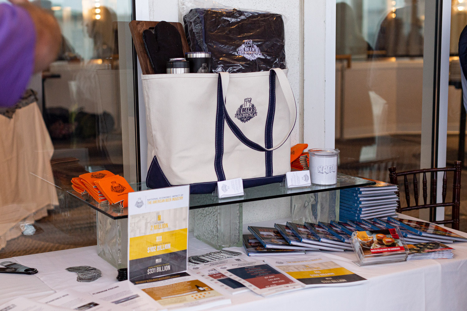 A table with a canvas tote bag with the Beer Institute logo, orange koozies, booklets and printed cards on a glass shelf.