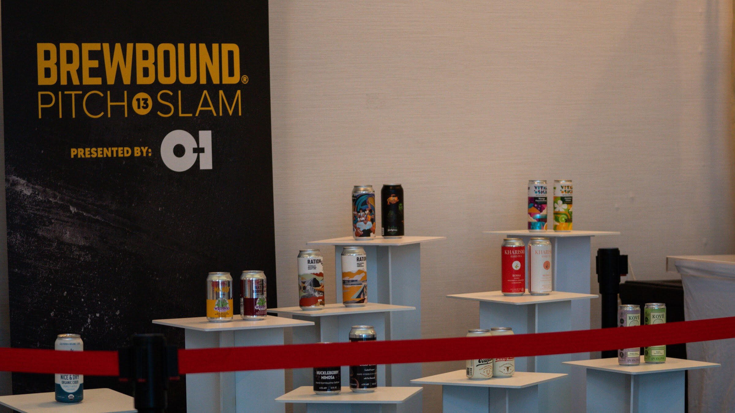 Cans arranged on white pedestals behind a red rope in front of a black Brewbound Pitch Slam banner presented by O-I.