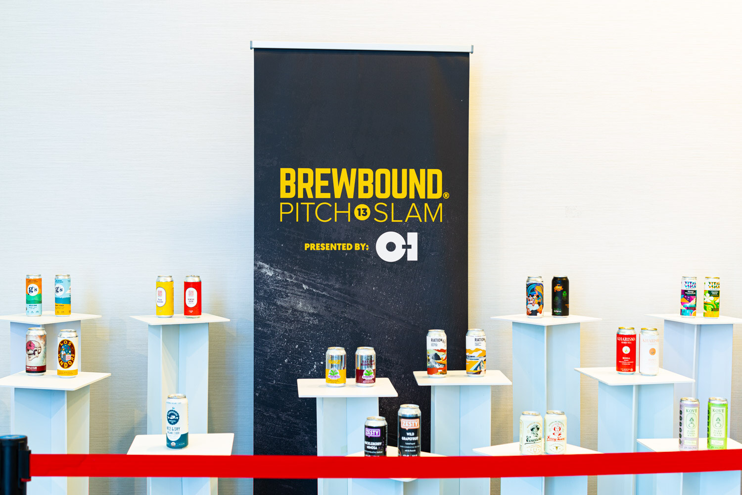 Cans on white pedestals behind a red rope in front of a black Brewbound Pitch Slam banner presented by O-I.