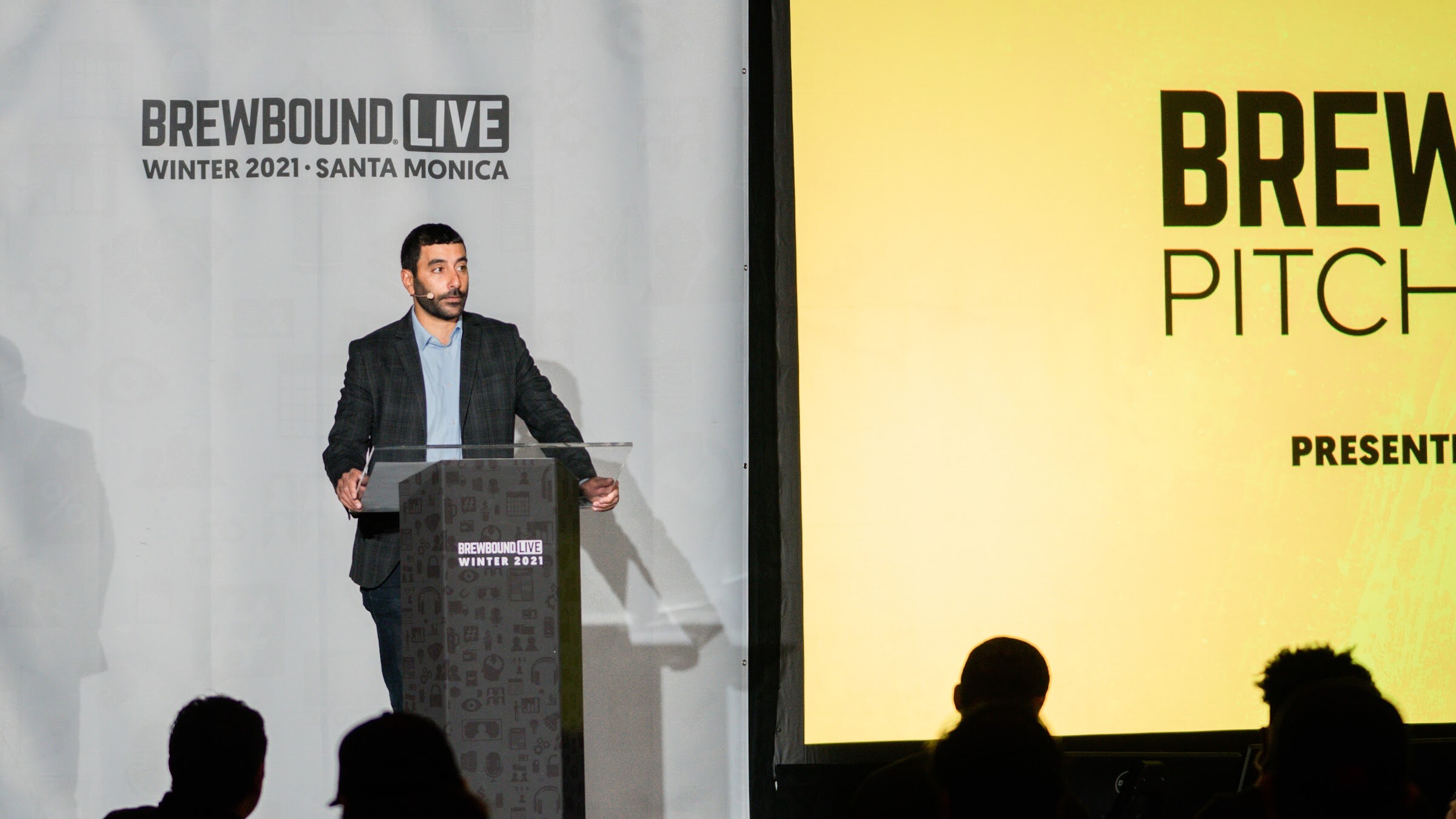 A man in a dark blazer stands at a clear podium in front of a yellow Brewbound Pitch Slam screen.