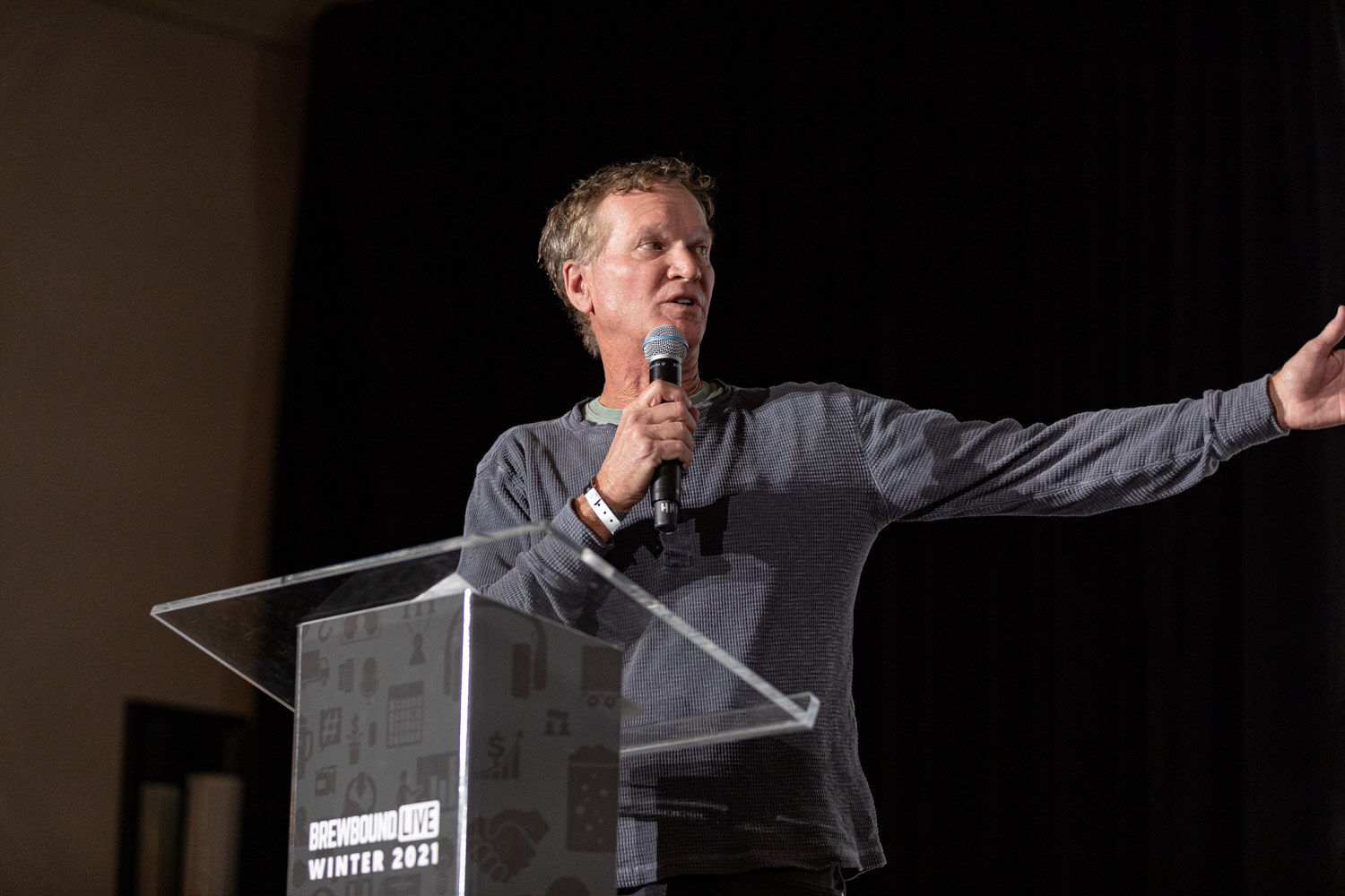A man in a gray sweater holds a microphone at the podium with one arm outstretched.