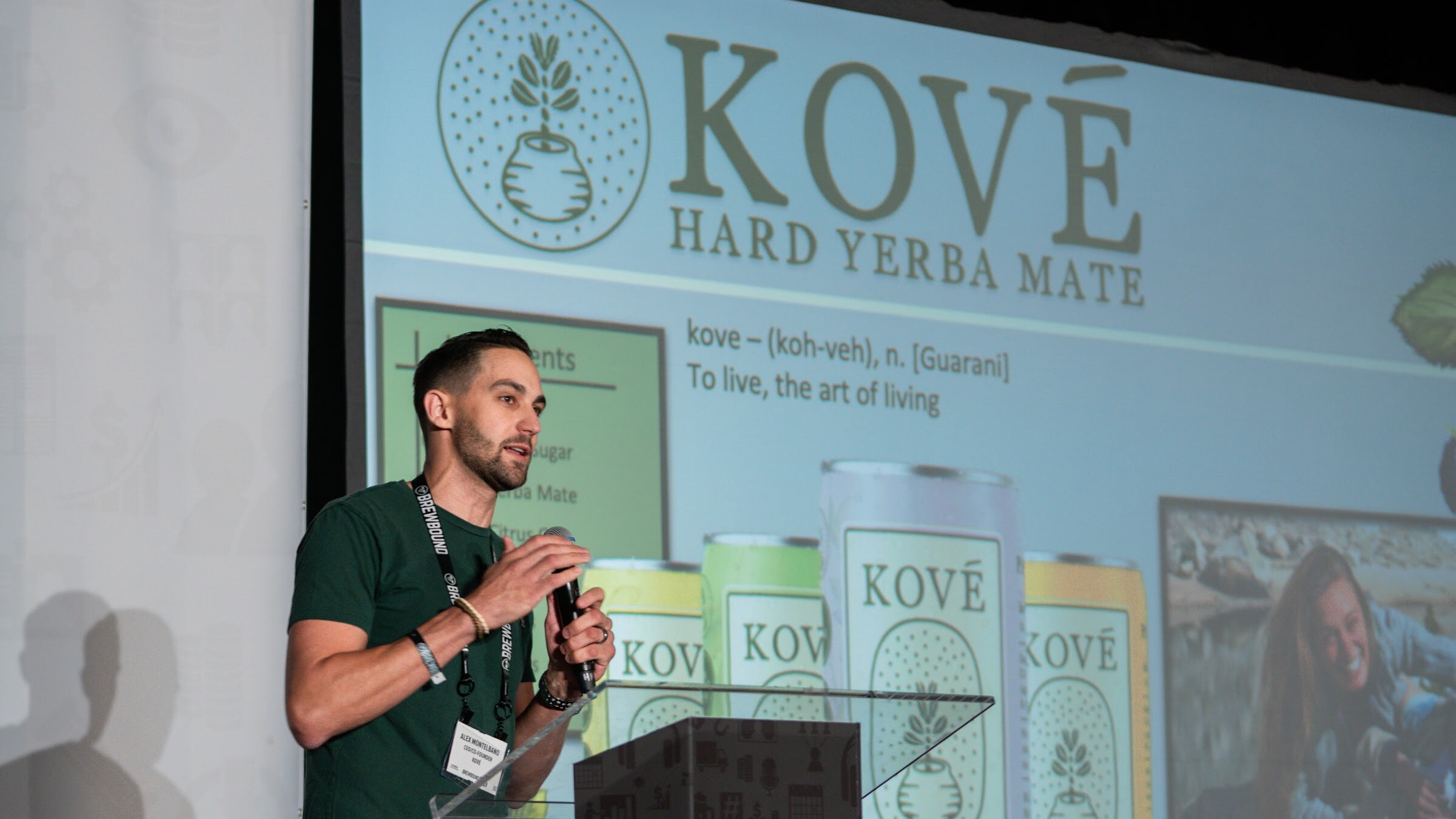 A man in a green shirt speaks into a microphone beside a projected slide for Kove hard yerba mate with cans of the product.