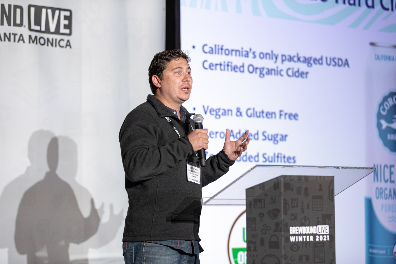 A man with a microphone speaks beside a slide listing organic cider attributes such as vegan and gluten free.