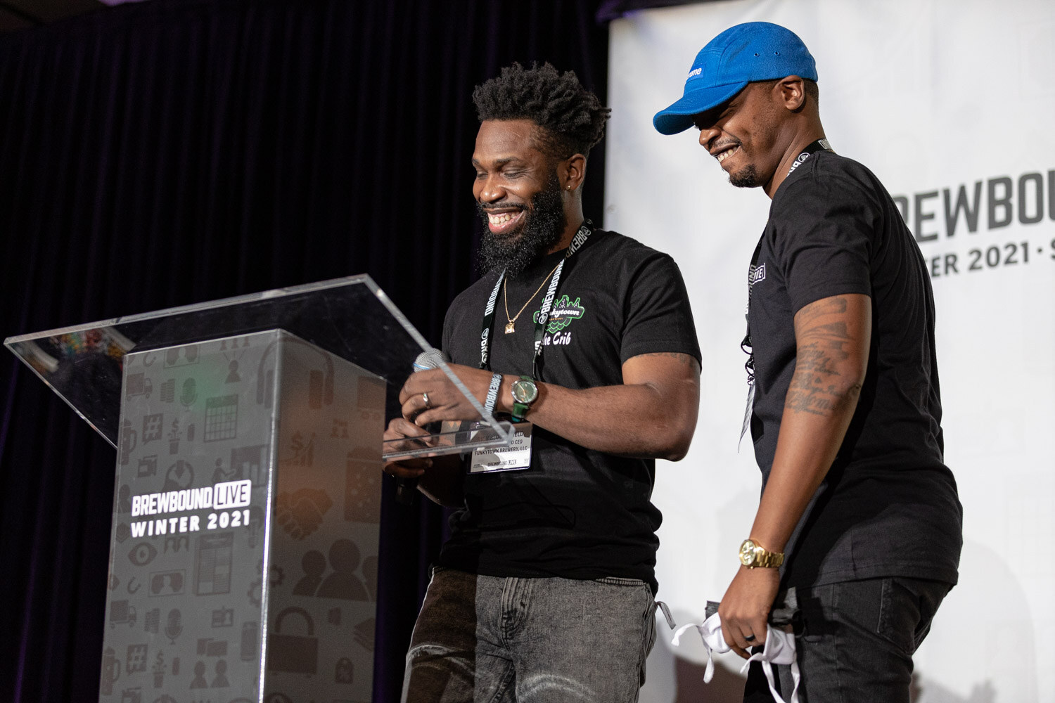 Two men in black shirts smile at a clear podium on stage.