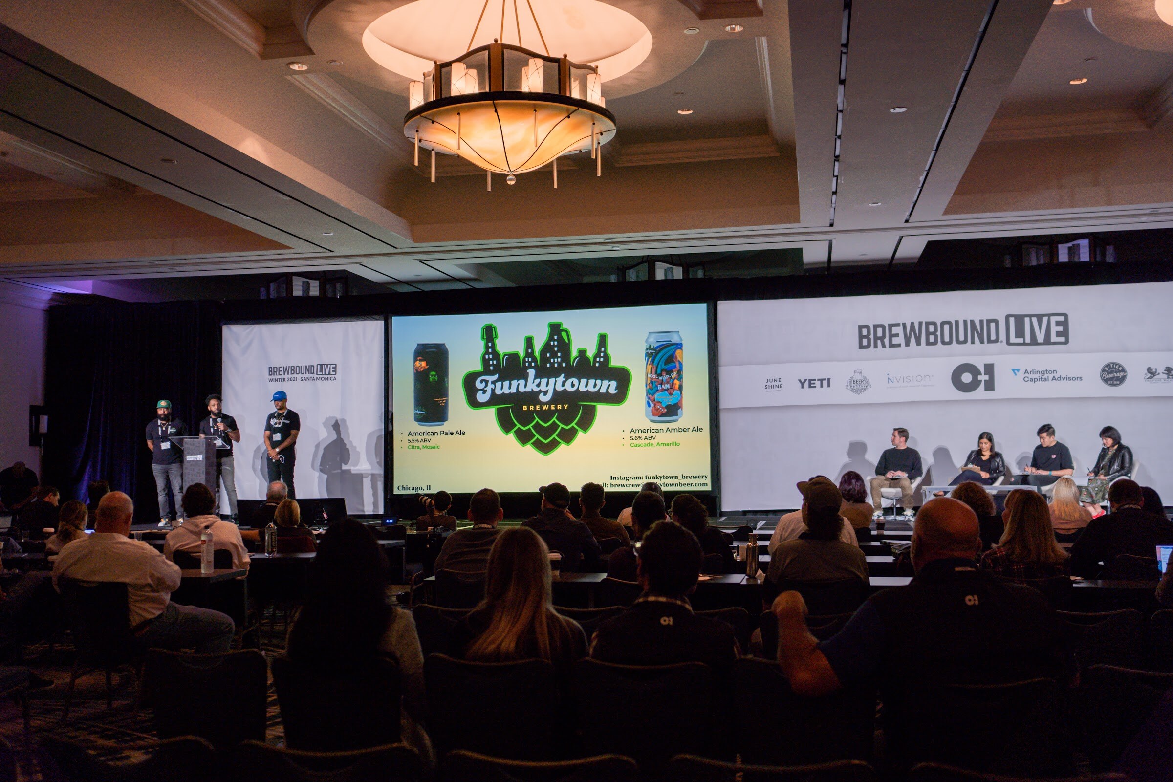 Wide view of a ballroom with three people on stage beside a screen showing the Funkytown logo, with judges seated at right and an audience in the foreground.