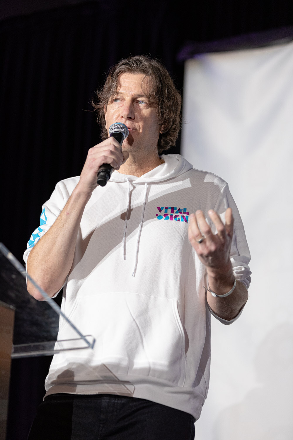 A man in a white hooded sweatshirt holds a microphone and gestures with one hand on a dark stage.