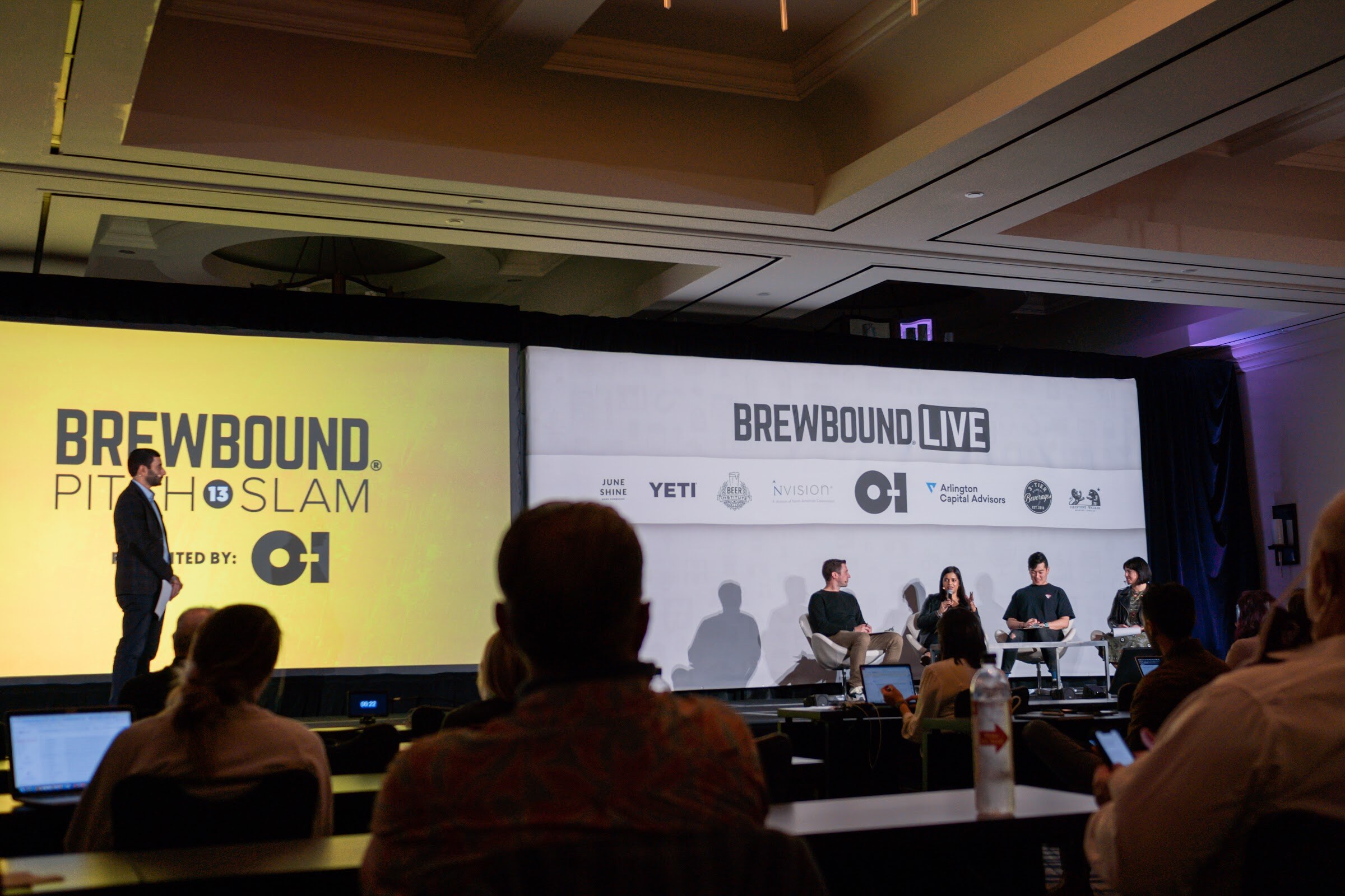 Wide view of a ballroom stage with a yellow Brewbound Pitch Slam screen, a man standing at left and four judges seated at right.