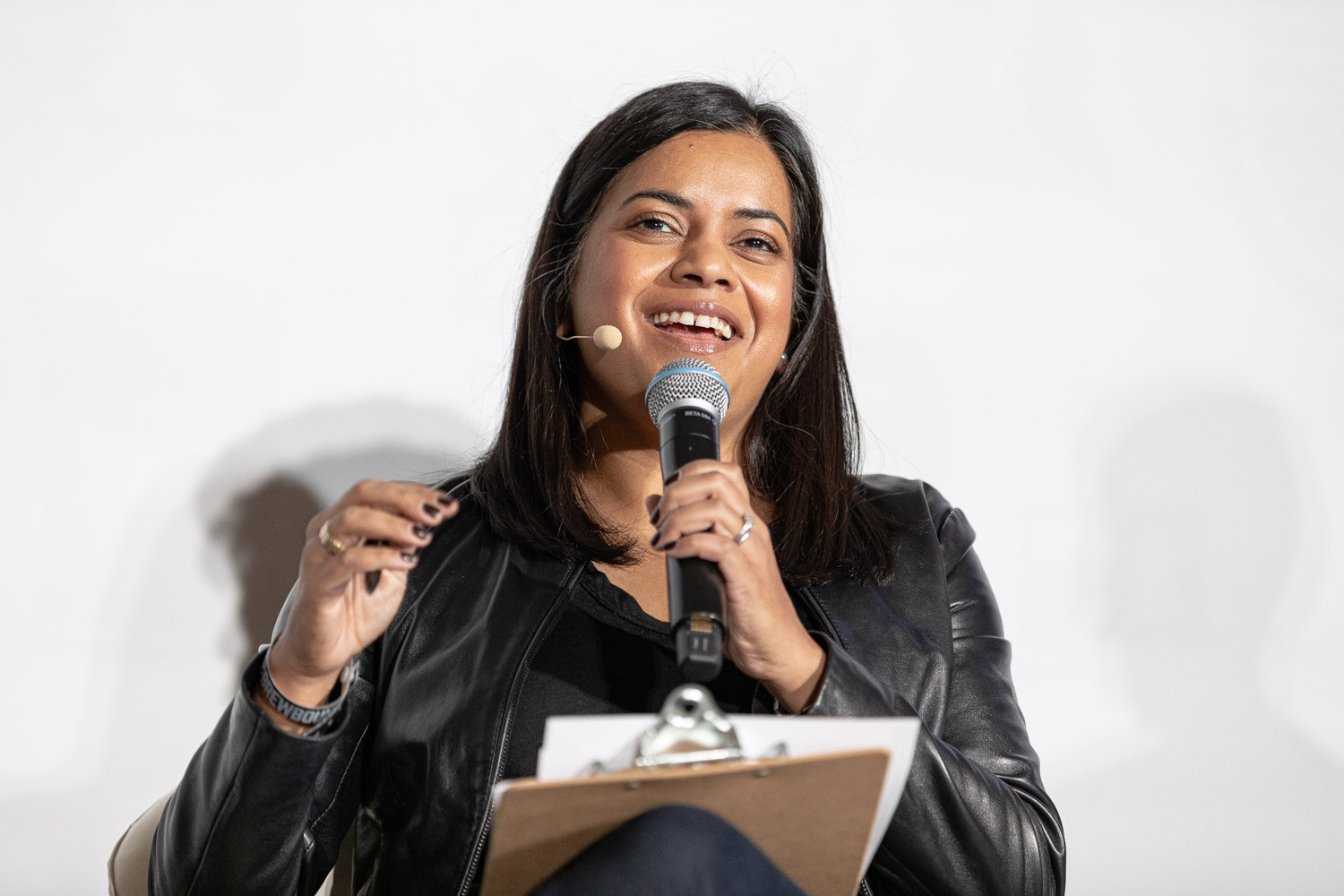 A smiling woman holding a microphone and clipboard speaks while seated on stage.