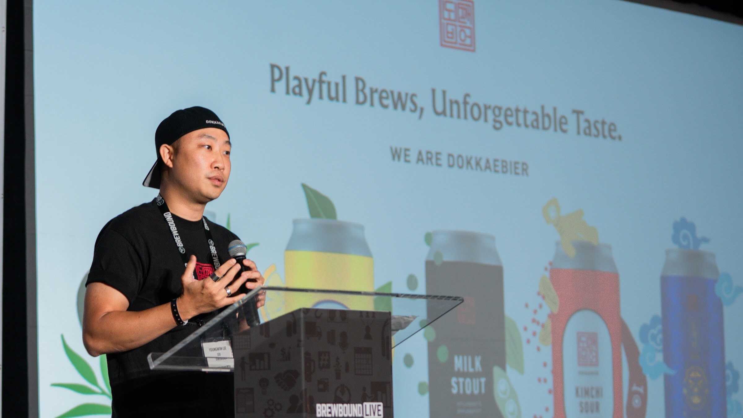 A man in a backwards cap speaks at the podium in front of a slide reading Playful Brews, Unforgettable Taste, We Are Dokkaebier.