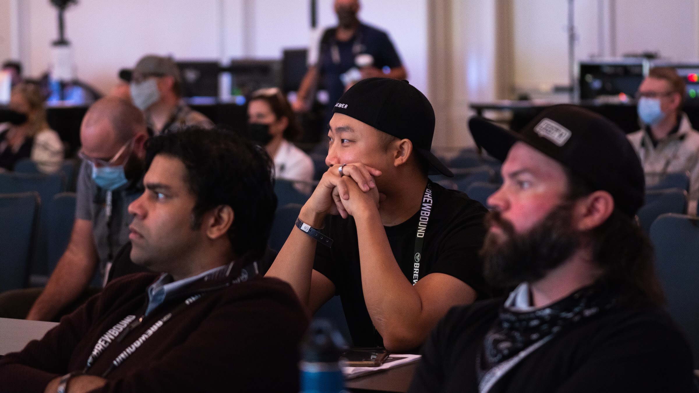 Three men in lanyards and caps sit in the audience looking toward the stage.