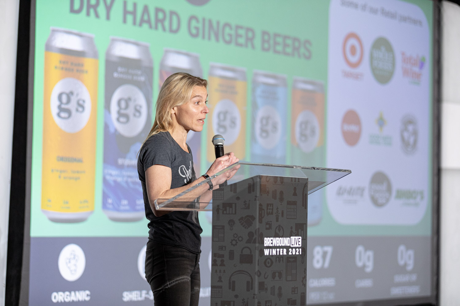 A woman speaks at the podium beside a slide showing cans of g's dry hard ginger beers and retail partner logos.